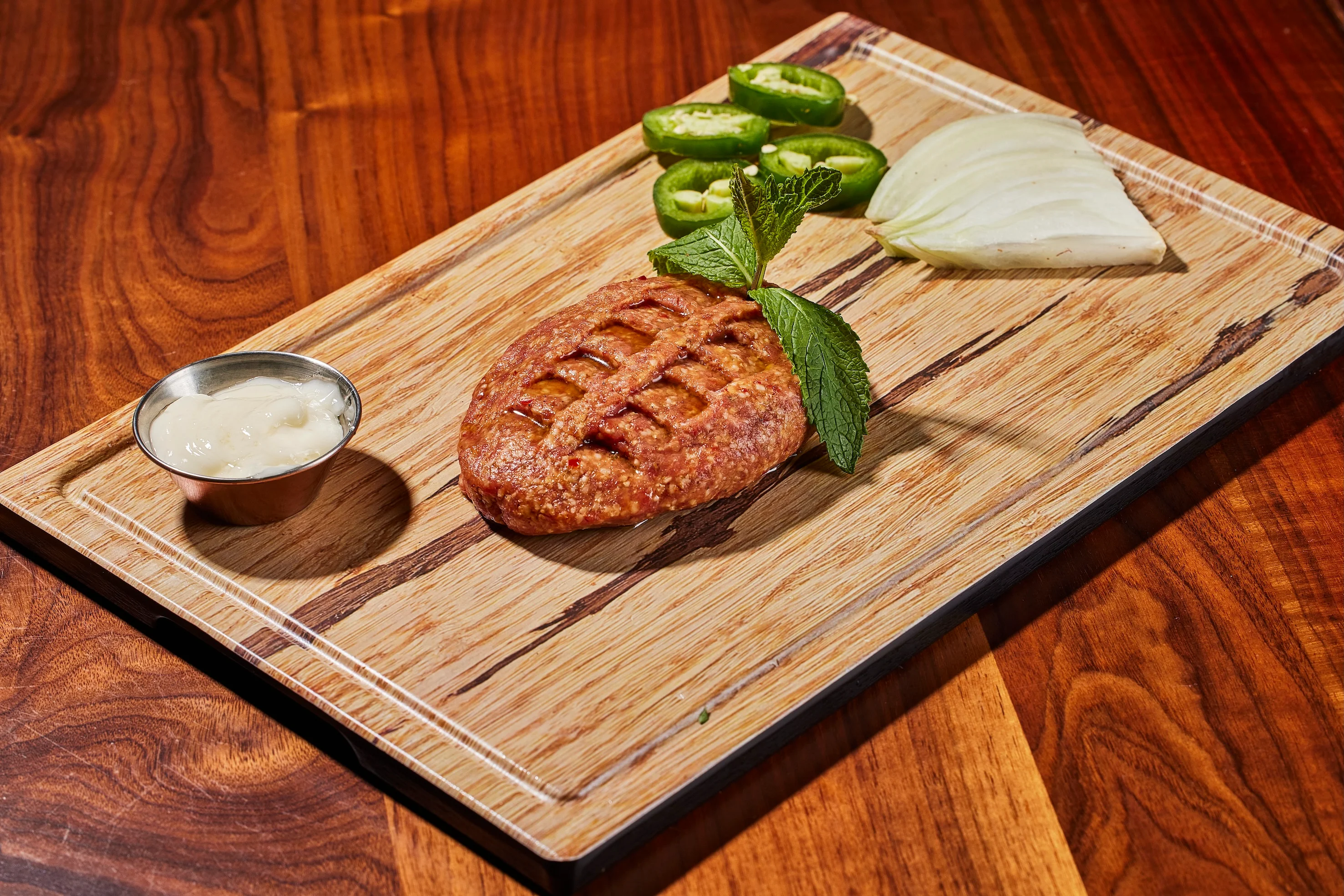 Grilled kebab with mint garnish on wooden board, alongside sliced jalapeños, onion wedge, and a small cup of creamy sauce. Warm, rustic atmosphere.