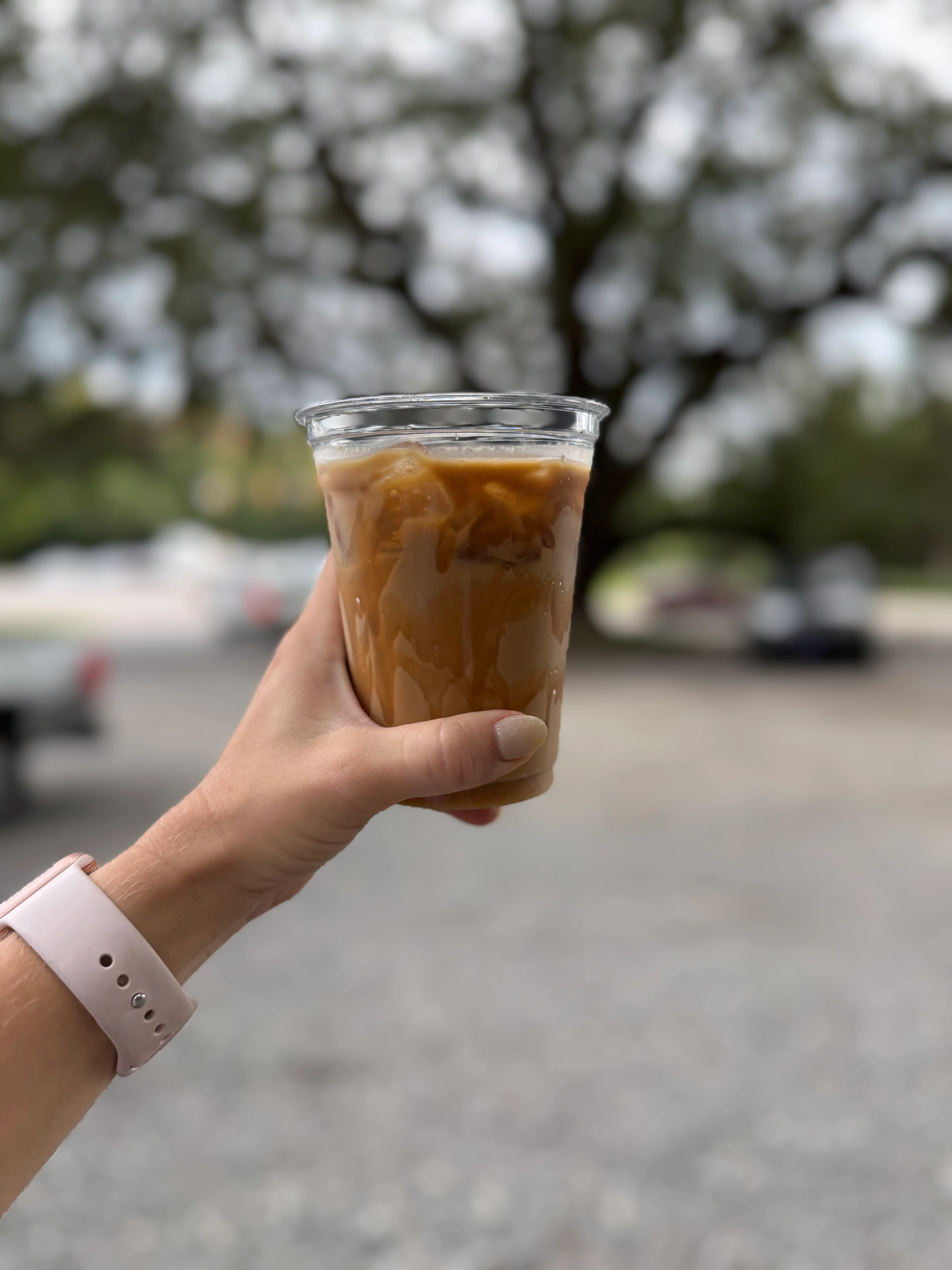 A hand holding a clear cup filled with iced coffee, with a blurred outdoor background featuring trees.