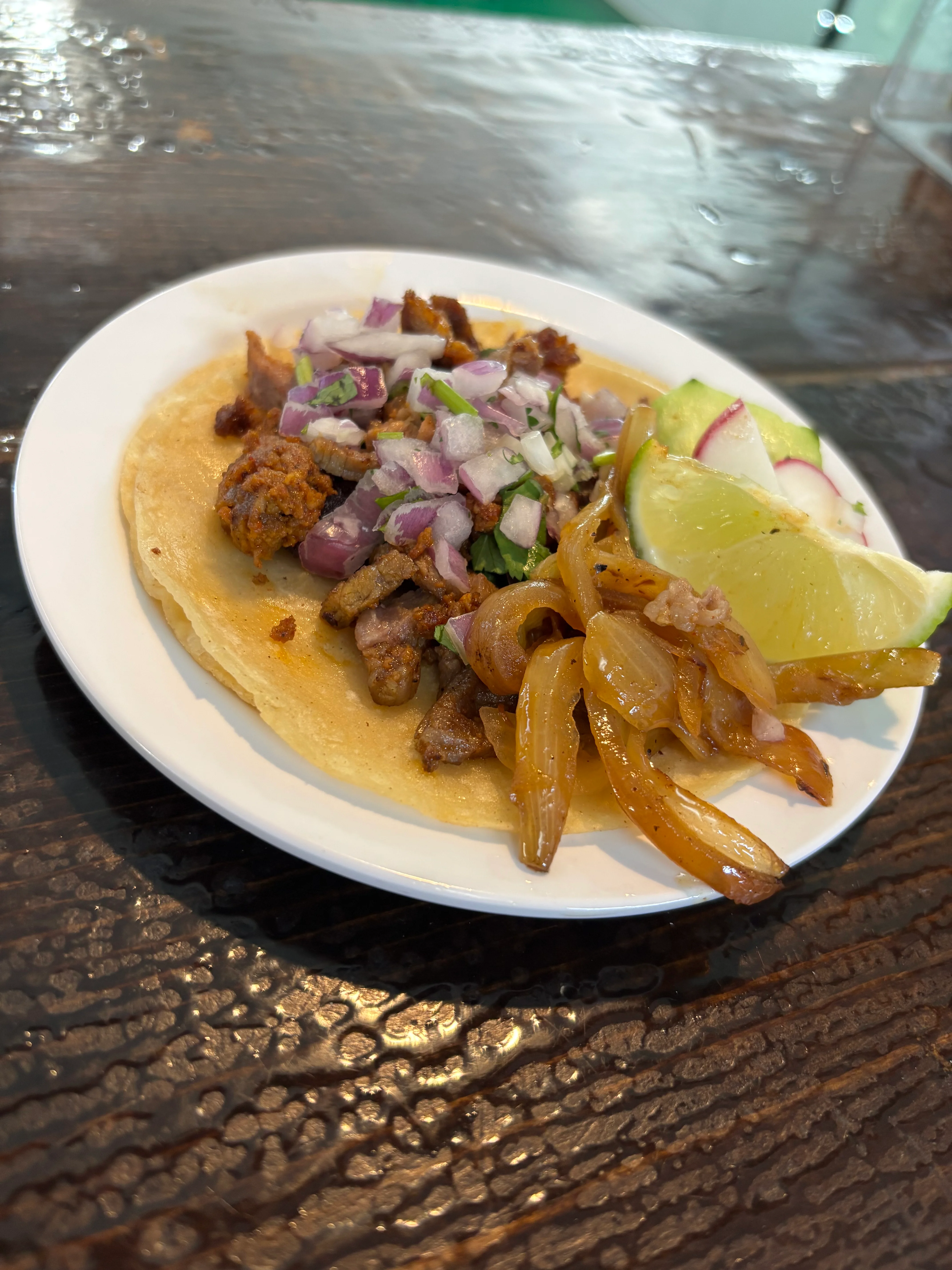 A serving of tacos served on a festive paper with a bowl of spicy sauce, lime wedges, and cucumber slices. The tacos are filled with meat and topped with cilantro and onions.