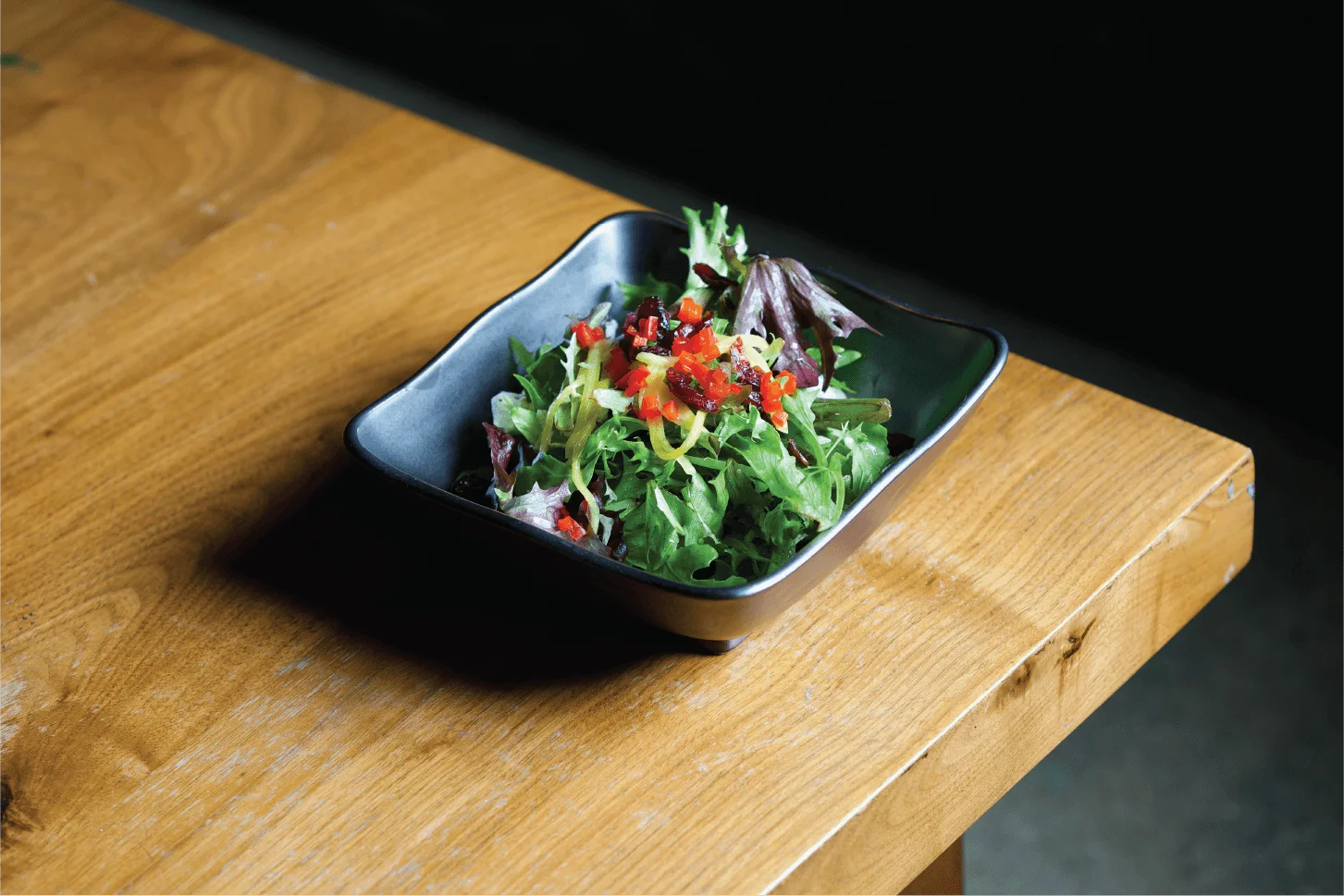 Fresh mixed greens salad with diced red peppers and yellow toppings served in a dark square bowl on a wooden table