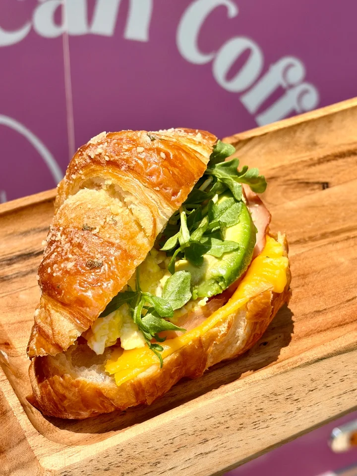 A croissant sandwich filled with avocado, cheese, egg, and greens, served on a wooden plate.