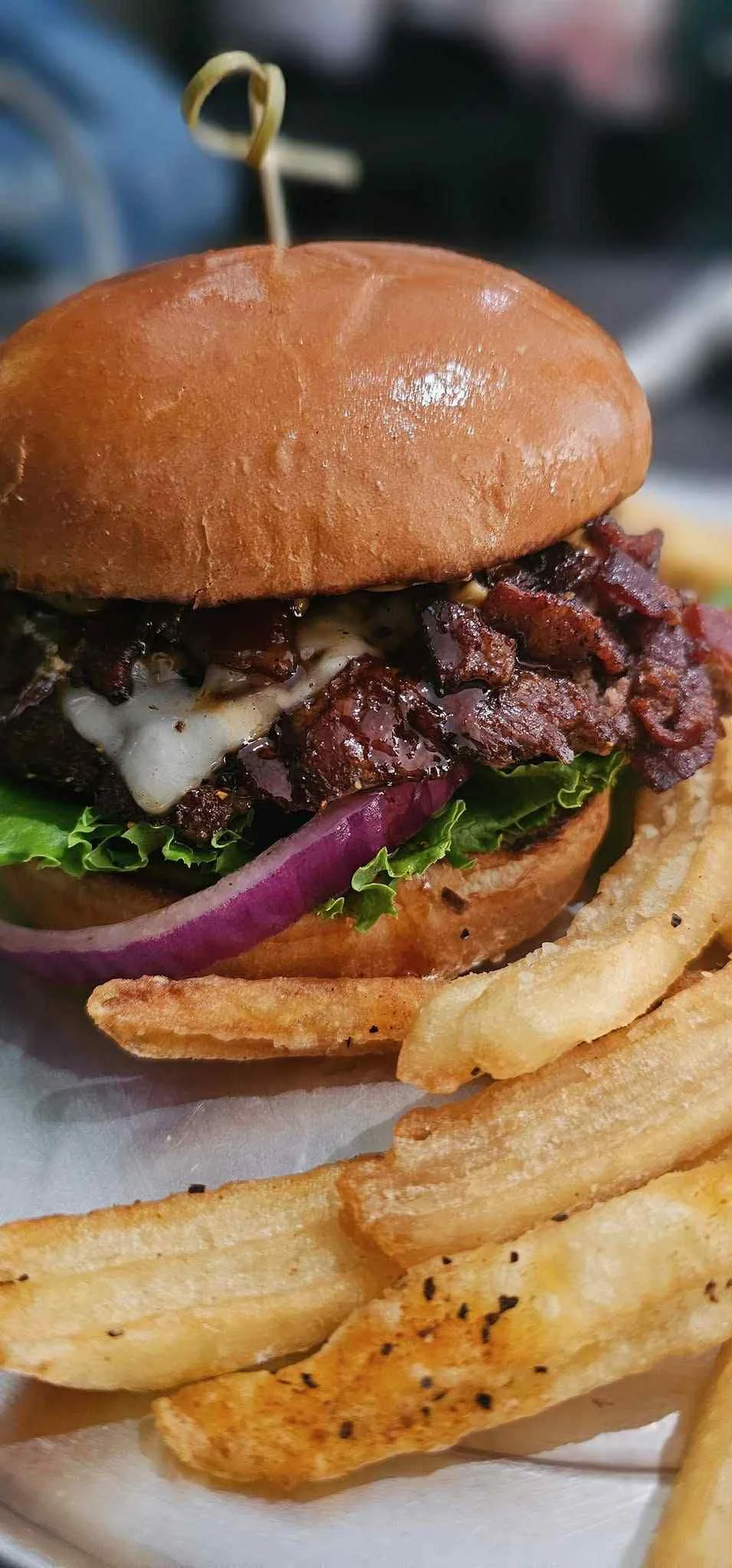 A close-up of a bacon cheeseburger with lettuce, onion, and melted cheese on a brioche bun, served with seasoned fries.