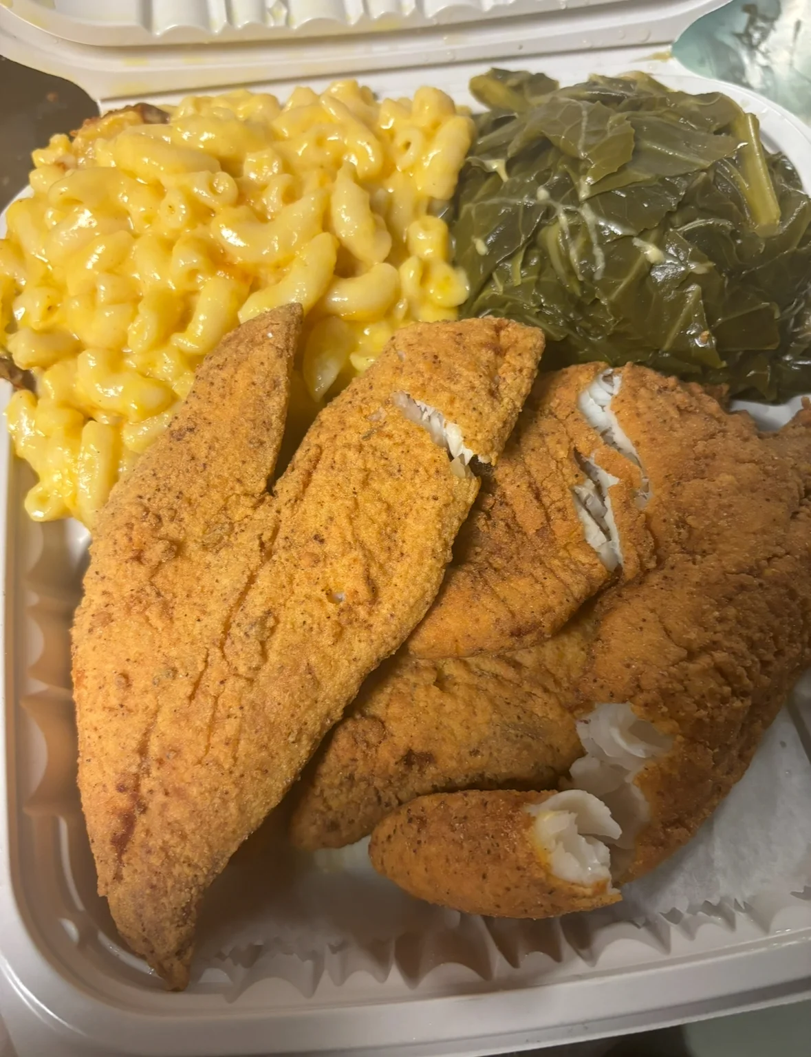 A plate featuring fried fish fillets with macaroni and cheese and steamed collard greens in a to-go box.