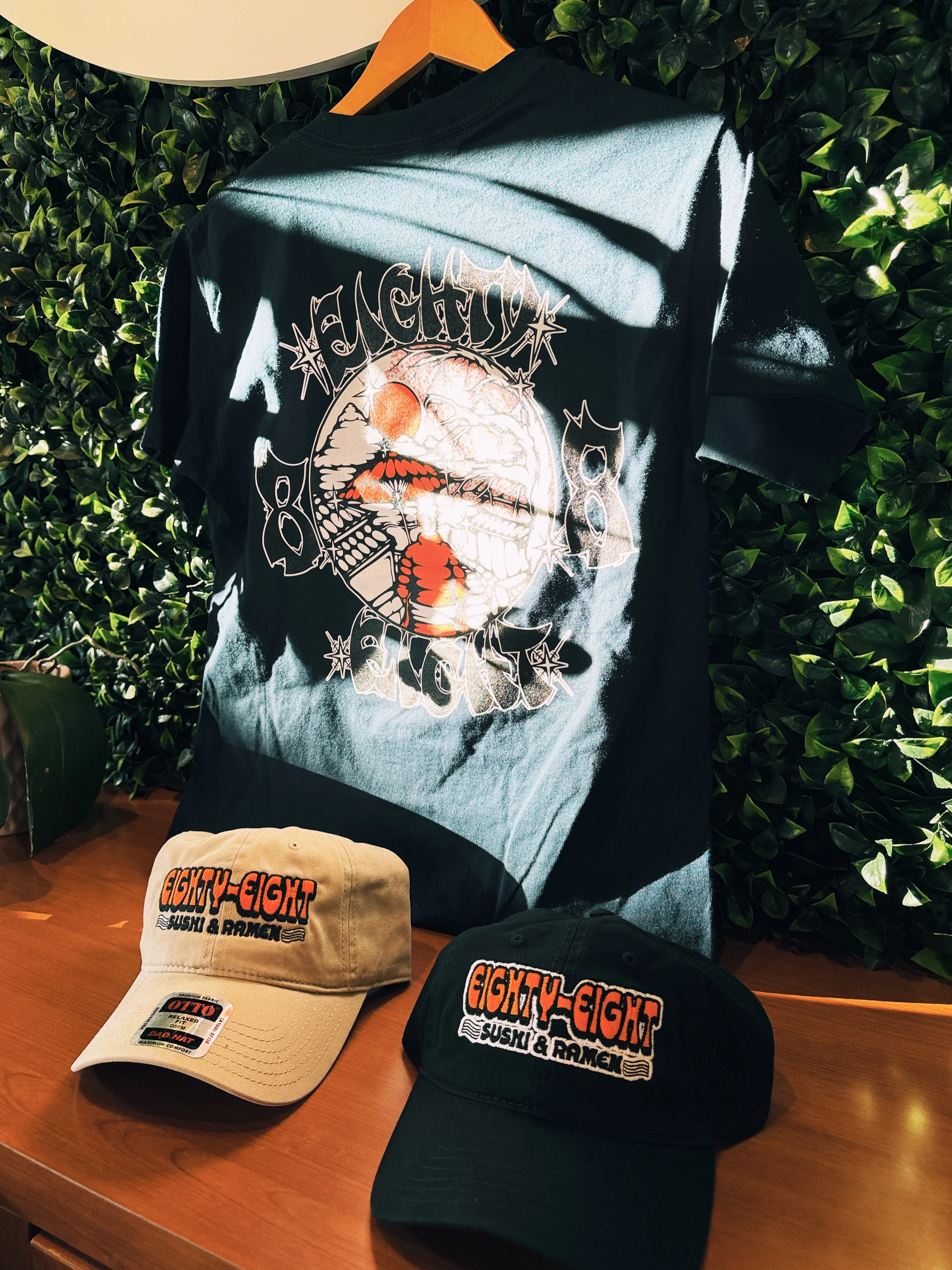 A black t-shirt displayed alongside two caps. The shirt features an artistic graphic design, while the caps display the logo for a sushi and ramen brand.