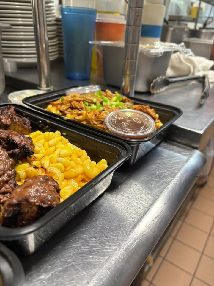 Two meal containers on a stainless steel surface, one filled with macaroni and cheese alongside grilled meat, the other with shredded meat and a small container of sauce.