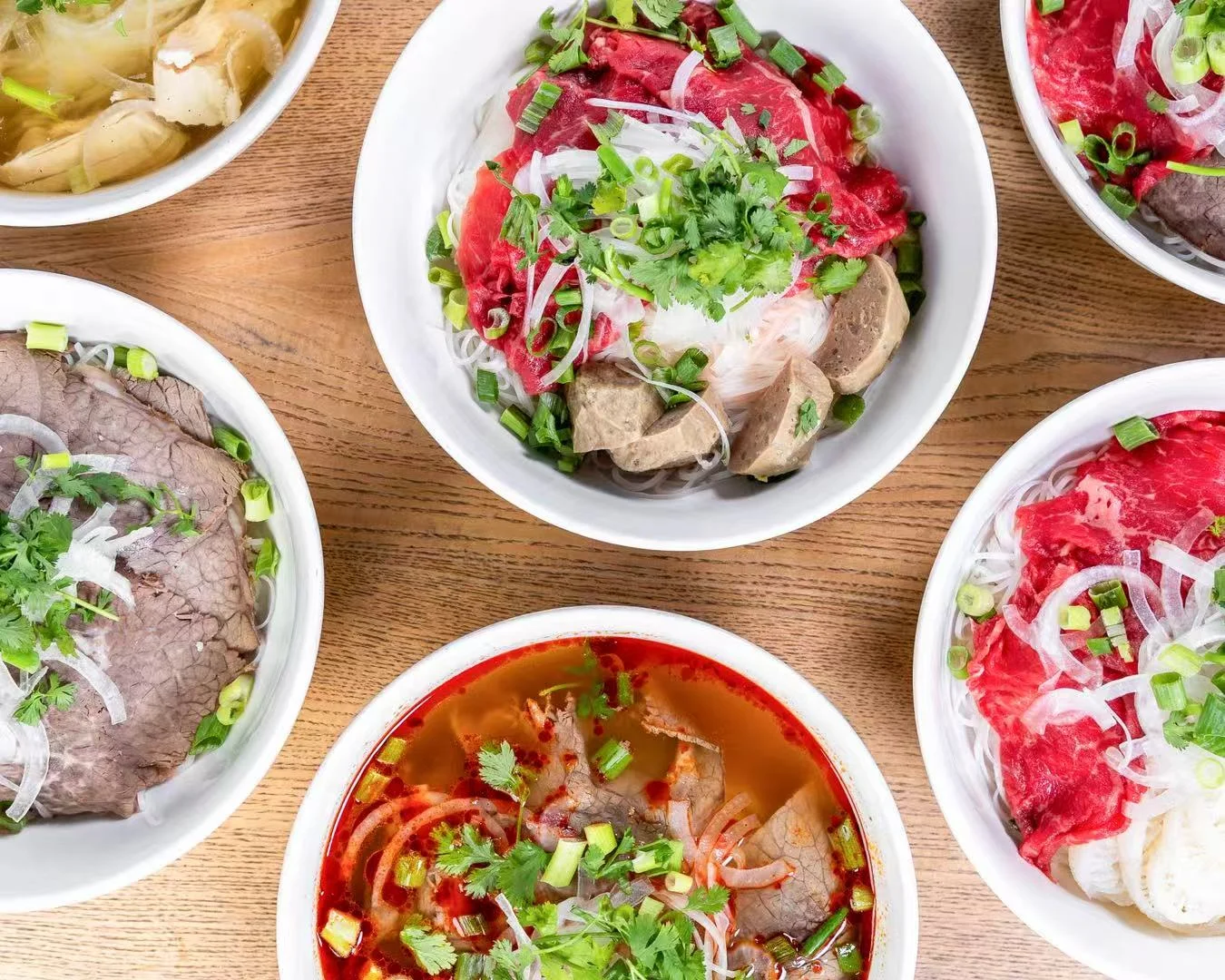 Bowls of pho with various meats and fresh herbs on a wooden table.