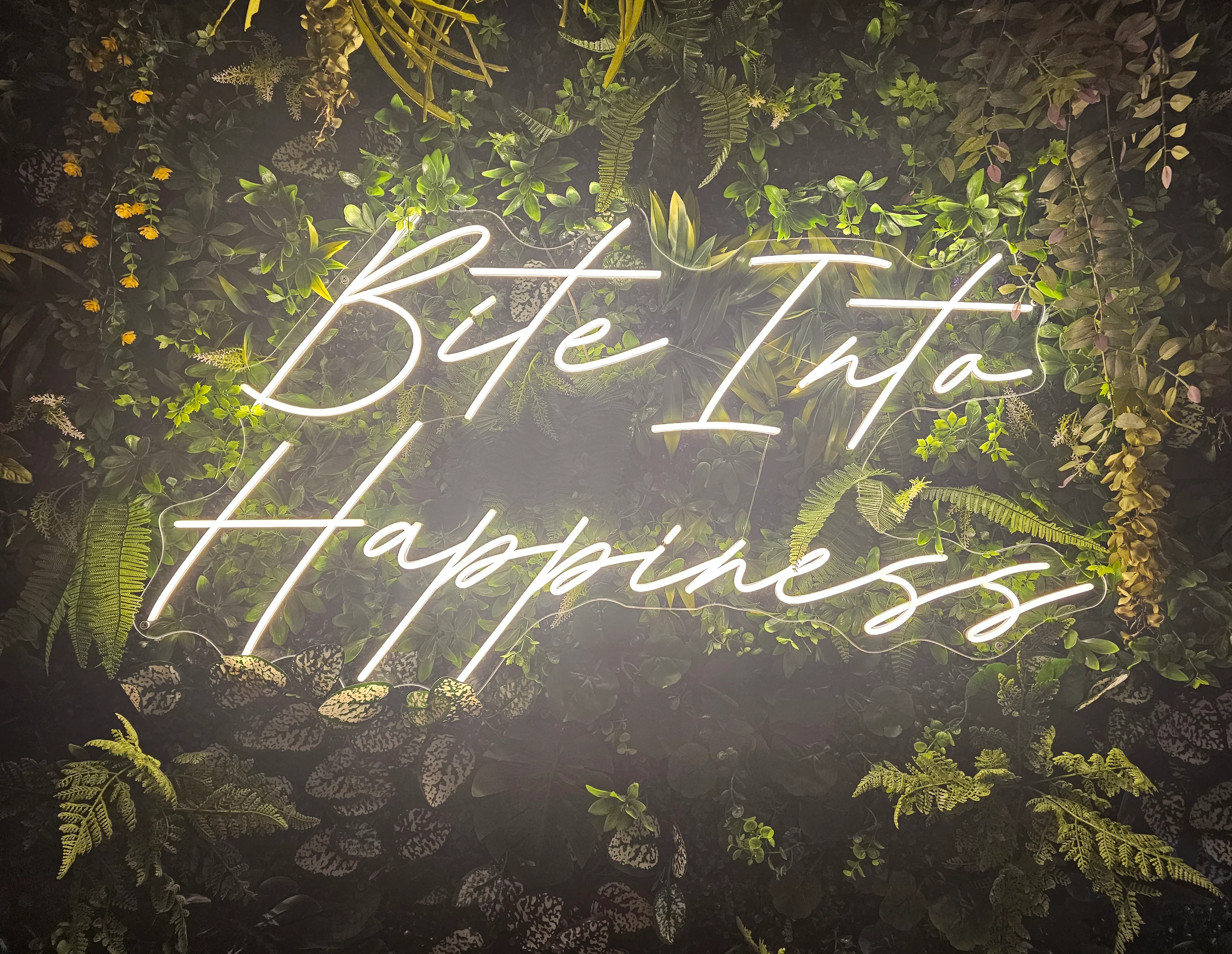 Neon sign reading 'Bite Into Happiness' surrounded by greenery
