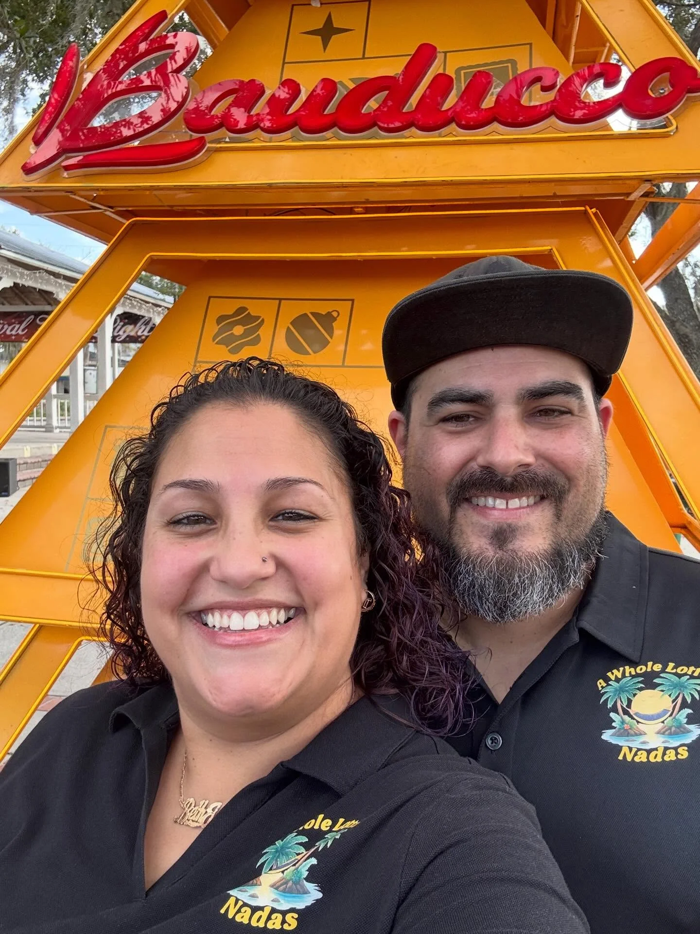 A cheerful couple posing for a photo in front of a colorful sign, both wearing matching shirts with a tropical design.
