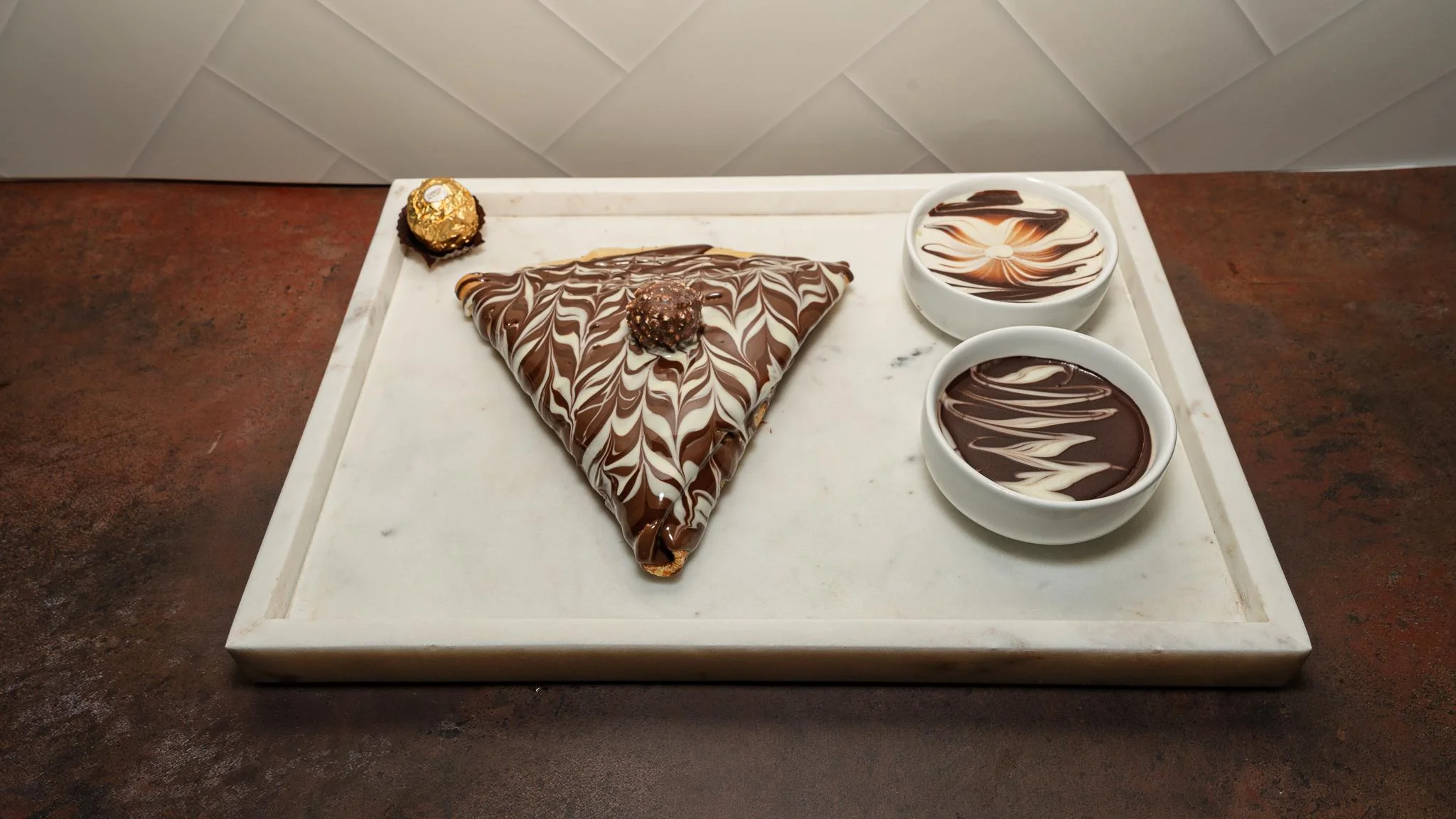 A luxury chocolate treat in a triangular form, finished with swirled chocolate, accompanied by two bowls of chocolate swirls on a marble tray.
