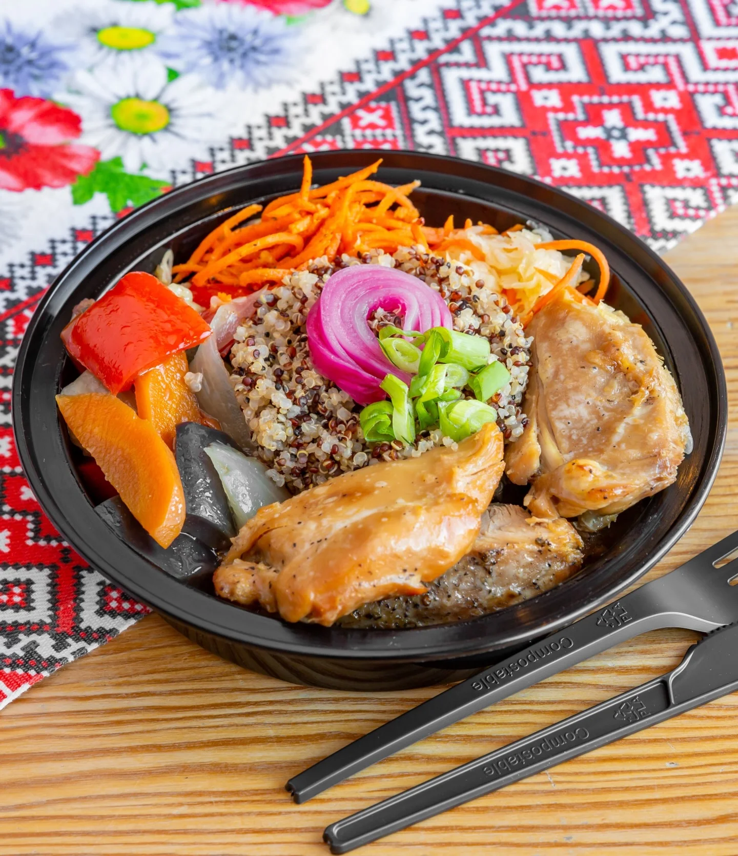 A bowl filled with quinoa topped with grilled chicken, vibrant vegetables including bell peppers, and garnished with green onions and pickled onions. The bowl is set against a colorful patterned background.