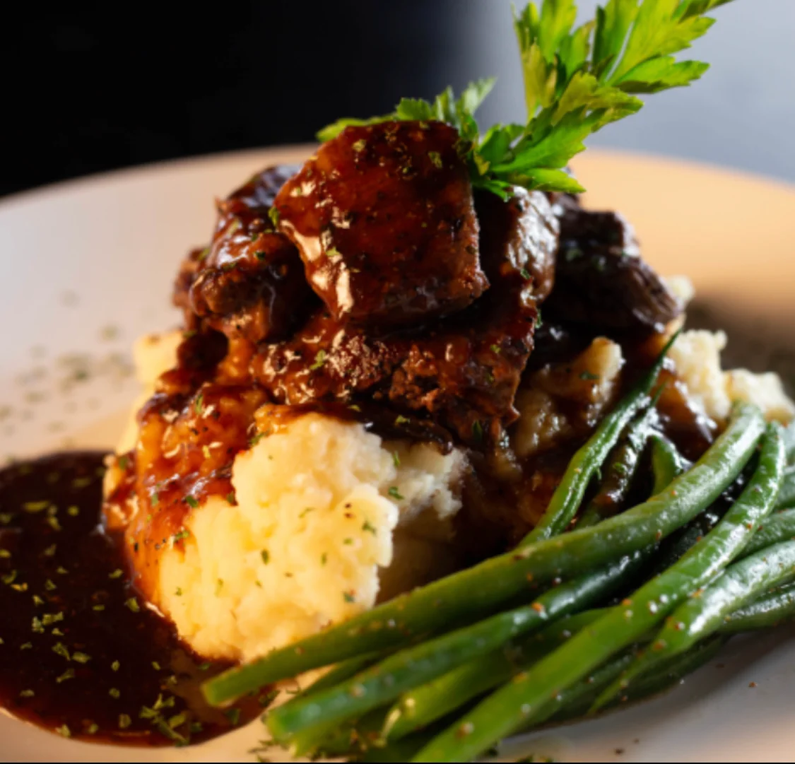 A serving of tender beef covered in a rich brown sauce atop creamy mashed potatoes, accompanied by green beans and garnished with a sprig of parsley.