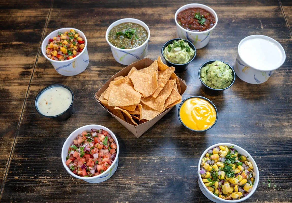 A variety of dips and sides including chips, salsa, guacamole, and sauces arranged around a wooden table.