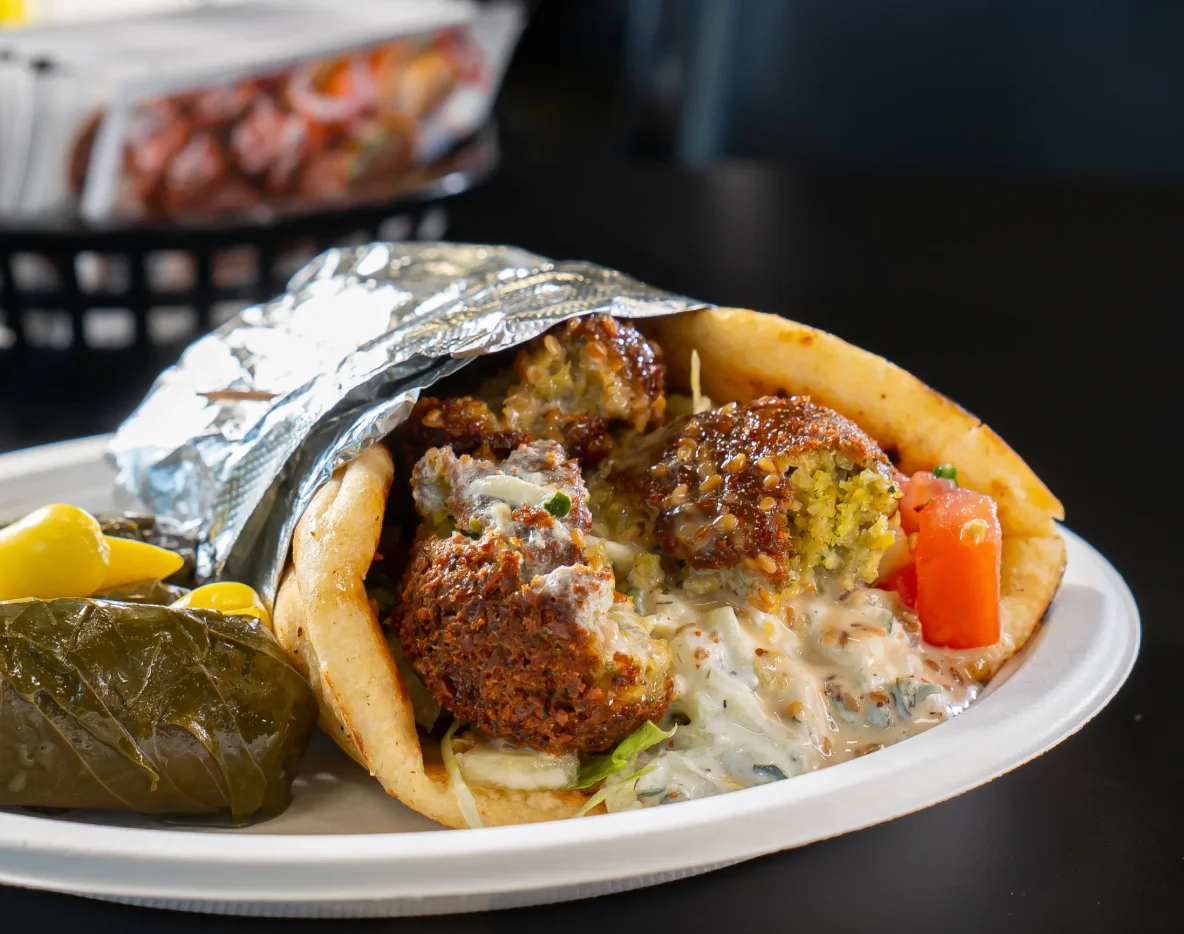 A wrap filled with falafel balls, fresh vegetables, and drizzled with sauce, served with a side of pickles and stuffed grape leaves.