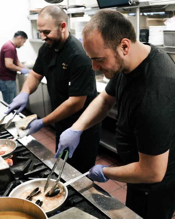 Two chefs preparing food in a professional kitchen. Both are using tongs to work with different ingredients while wearing gloves.