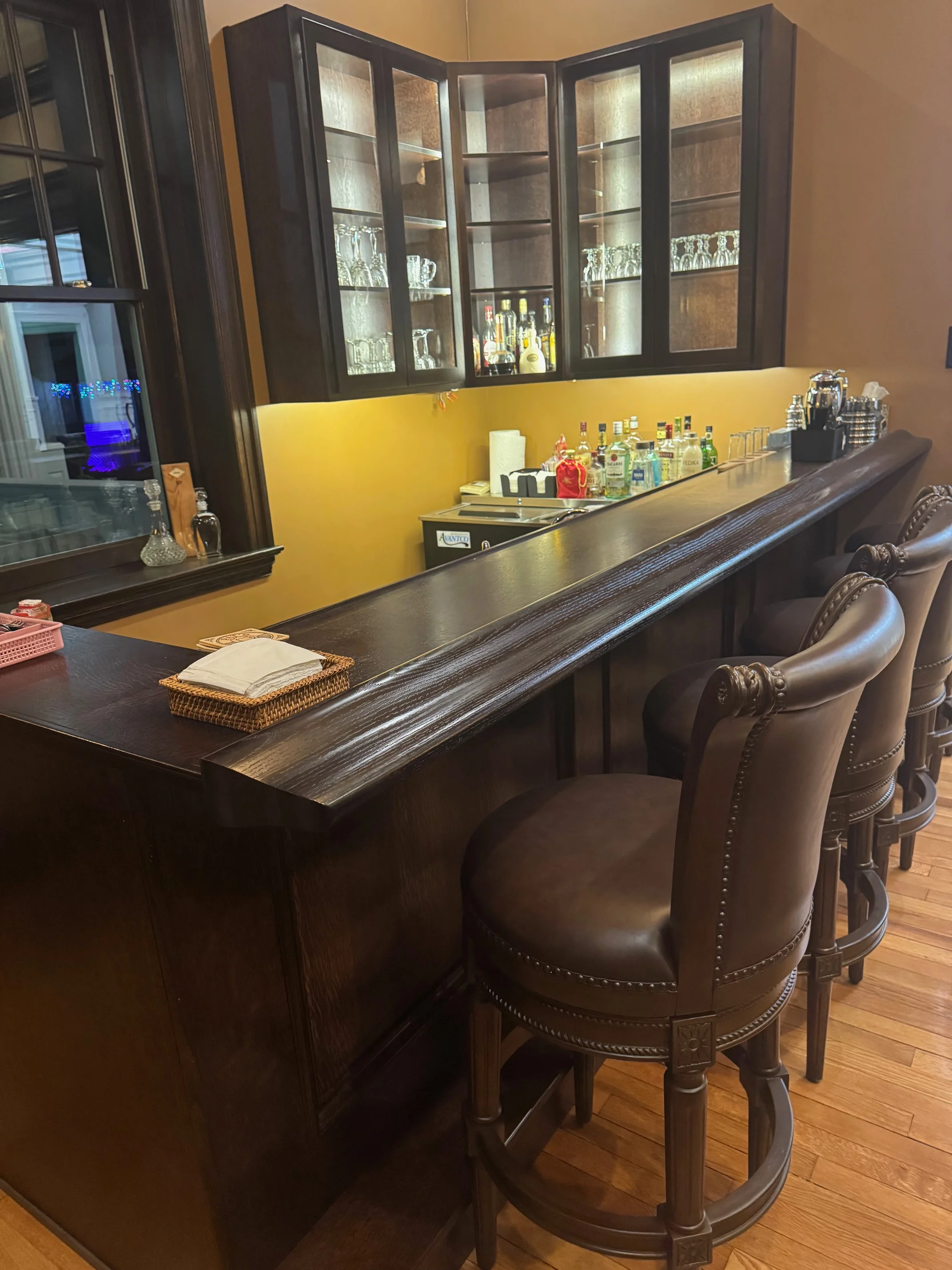 An elegant home bar with a polished wooden counter, surrounded by tall bar stools. Behind the bar, shelves display various bottles and glasses.