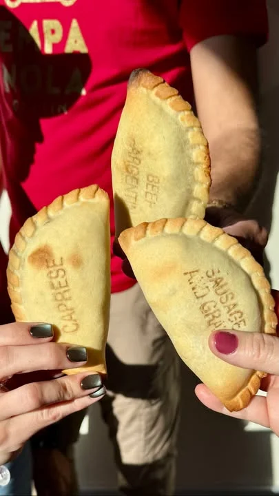 Three freshly baked empanadas held between hands, each featuring a different filling indicated by engraved names.