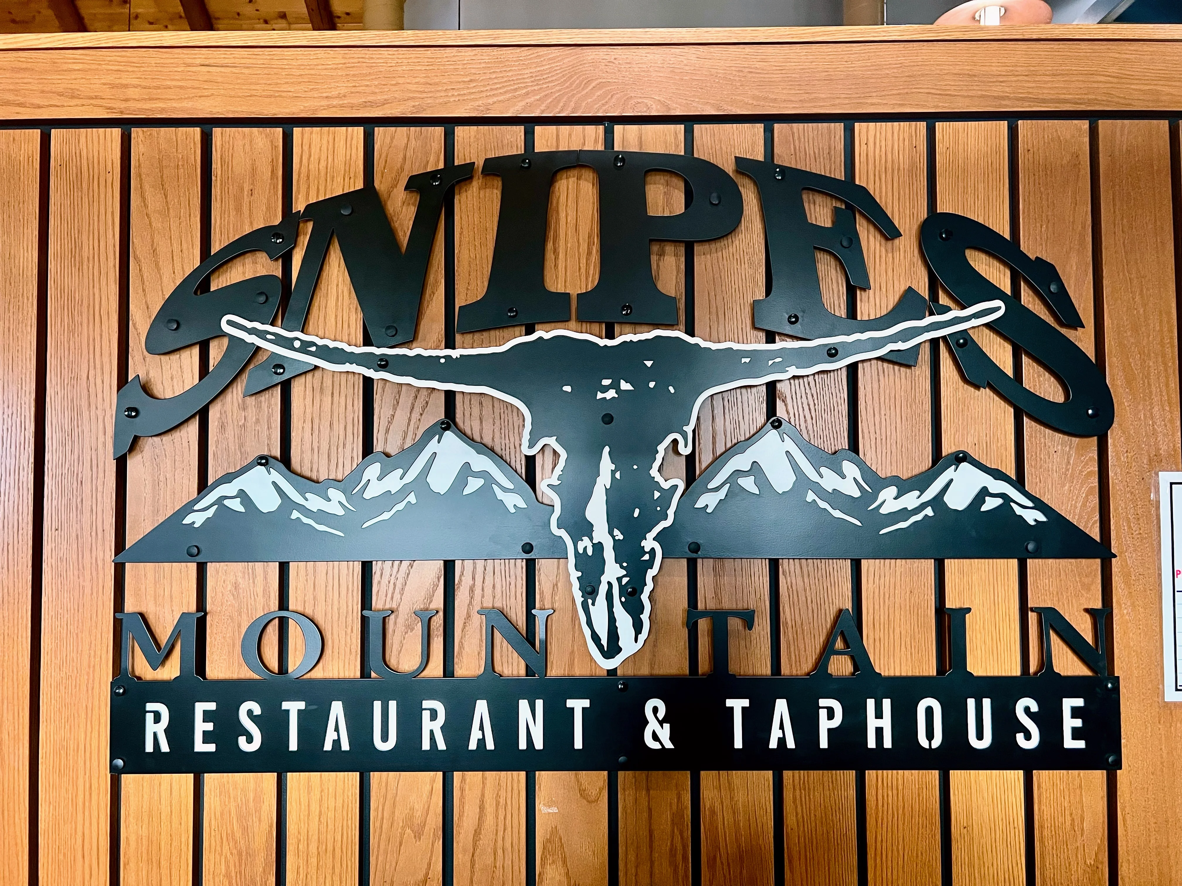 A decorative sign for a restaurant, featuring a longhorn skull and mountain graphics against a wood-paneled background.