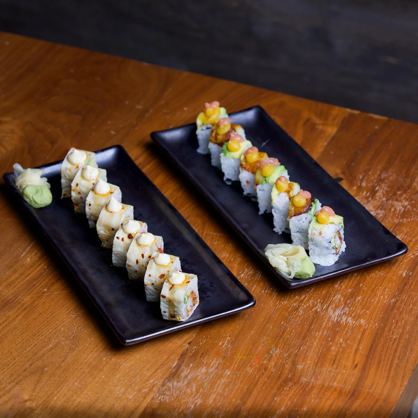 Two rectangular black plates on a wooden table holding different styles of sushi rolls, each garnished with colorful sauces and served with sides of pickled ginger and wasabi.