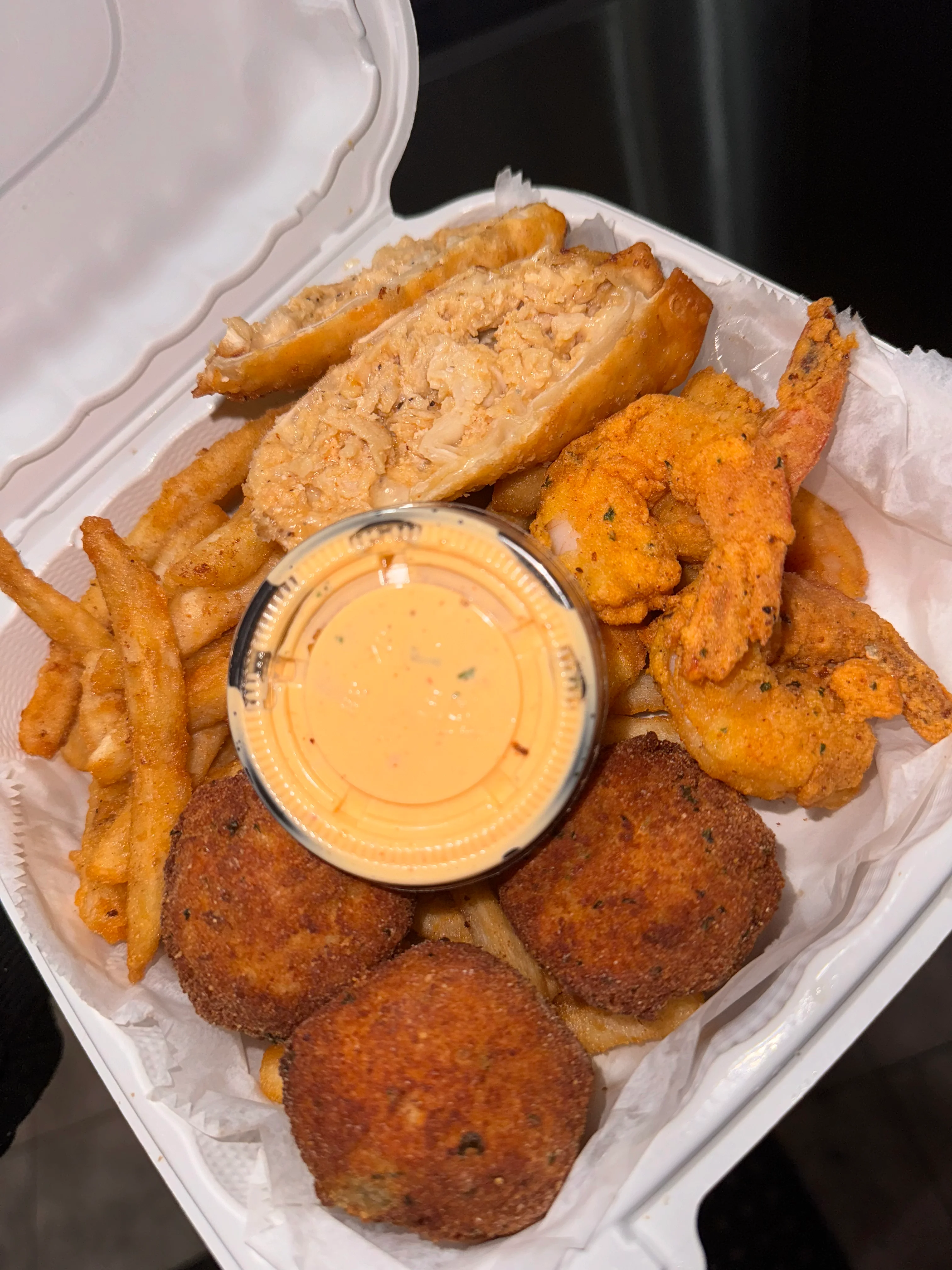 An assortment of fried seafood, chicken strips, French fries, and dipping sauce in a takeout container.
