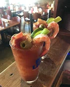 Two tall glasses of a cocktail garnished with shrimp, olives, and celery, sitting on a wooden bar.