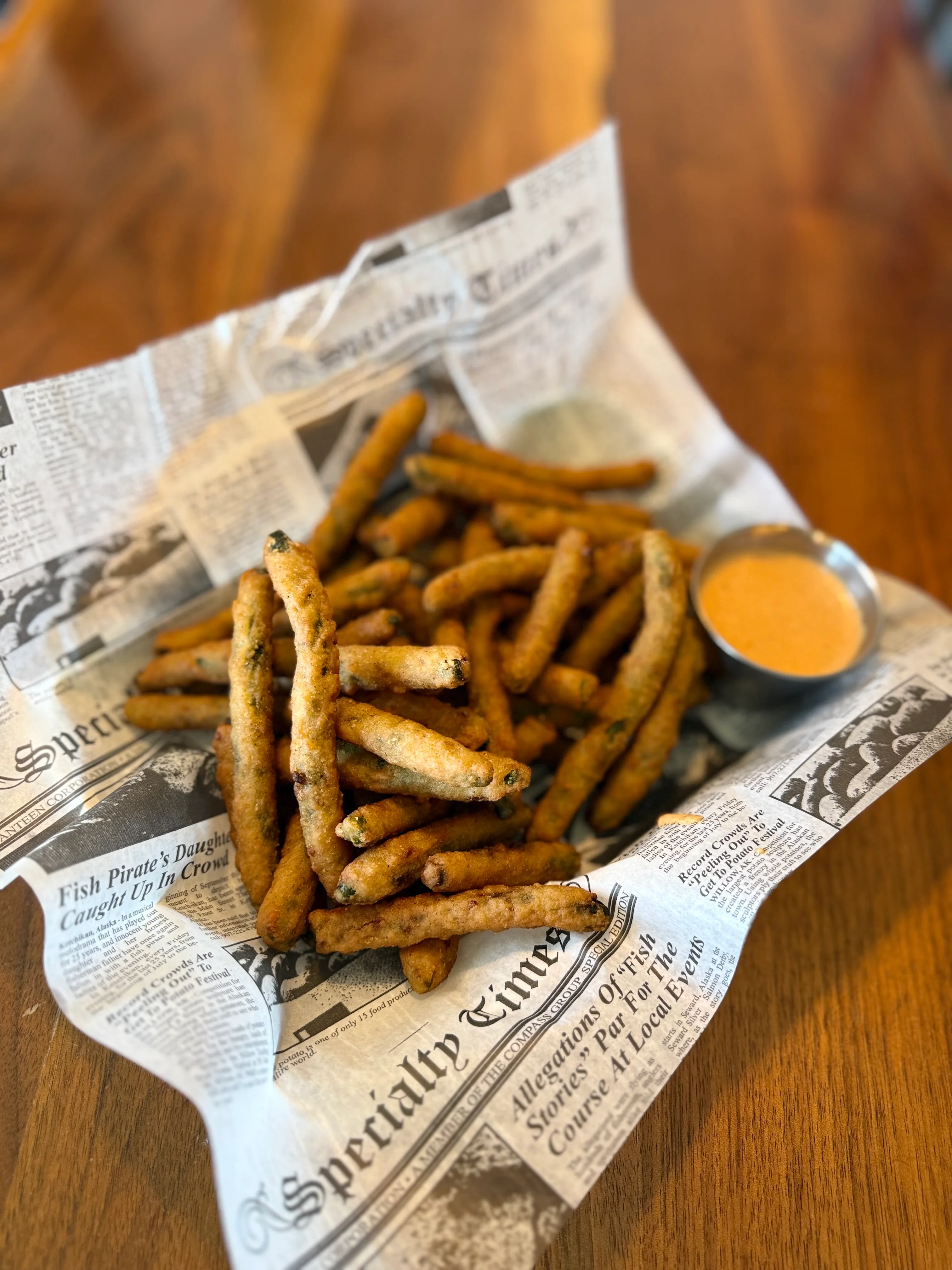 A plate of crispy fried pickles served with a dipping sauce, arranged in a basket lined with newspaper print.