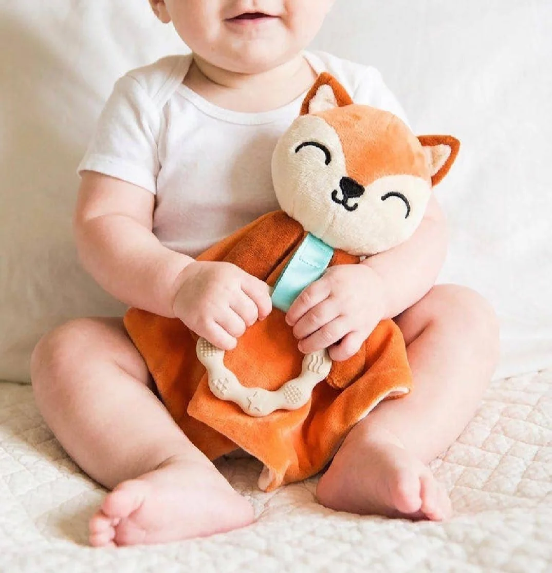 A baby sitting happily while holding a plush toy fox and a teething ring. The setting is cozy and warmth is conveyed through the soft colors.