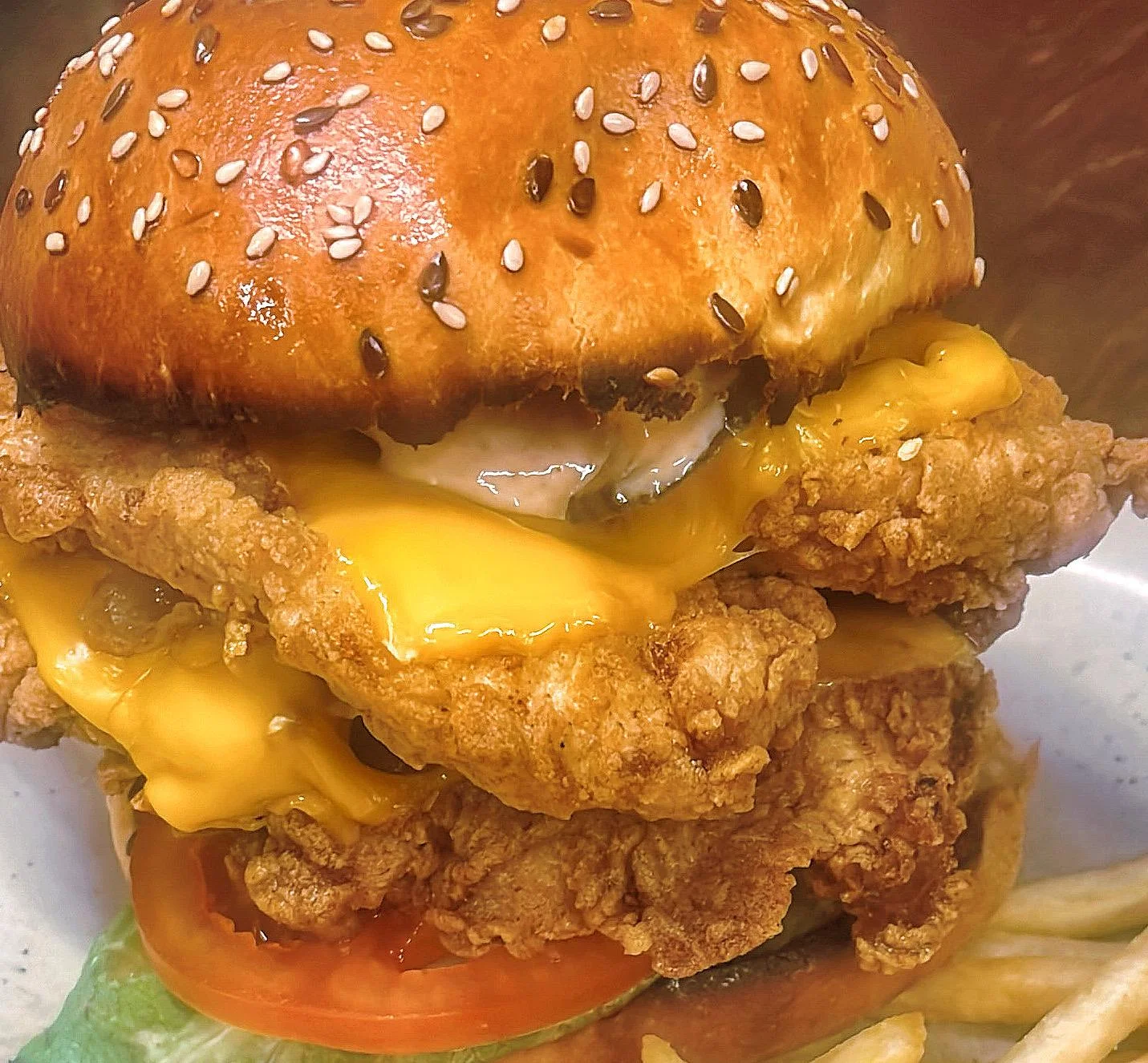 A towering chicken sandwich featuring crispy fried chicken, melted cheese, and fresh tomato, all nestled between a sesame seed bun. A side of french fries is visible.