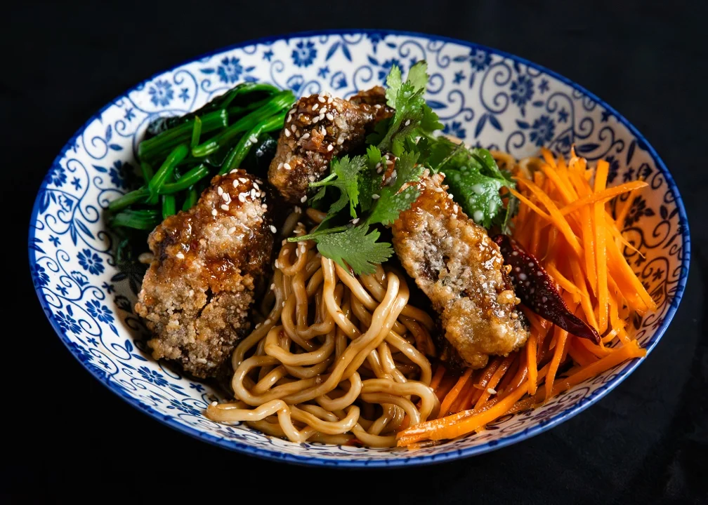 A beautifully arranged bowl of noodles topped with crispy pieces, sautéed greens, and shredded carrots, garnished with fresh herbs and sesame seeds.