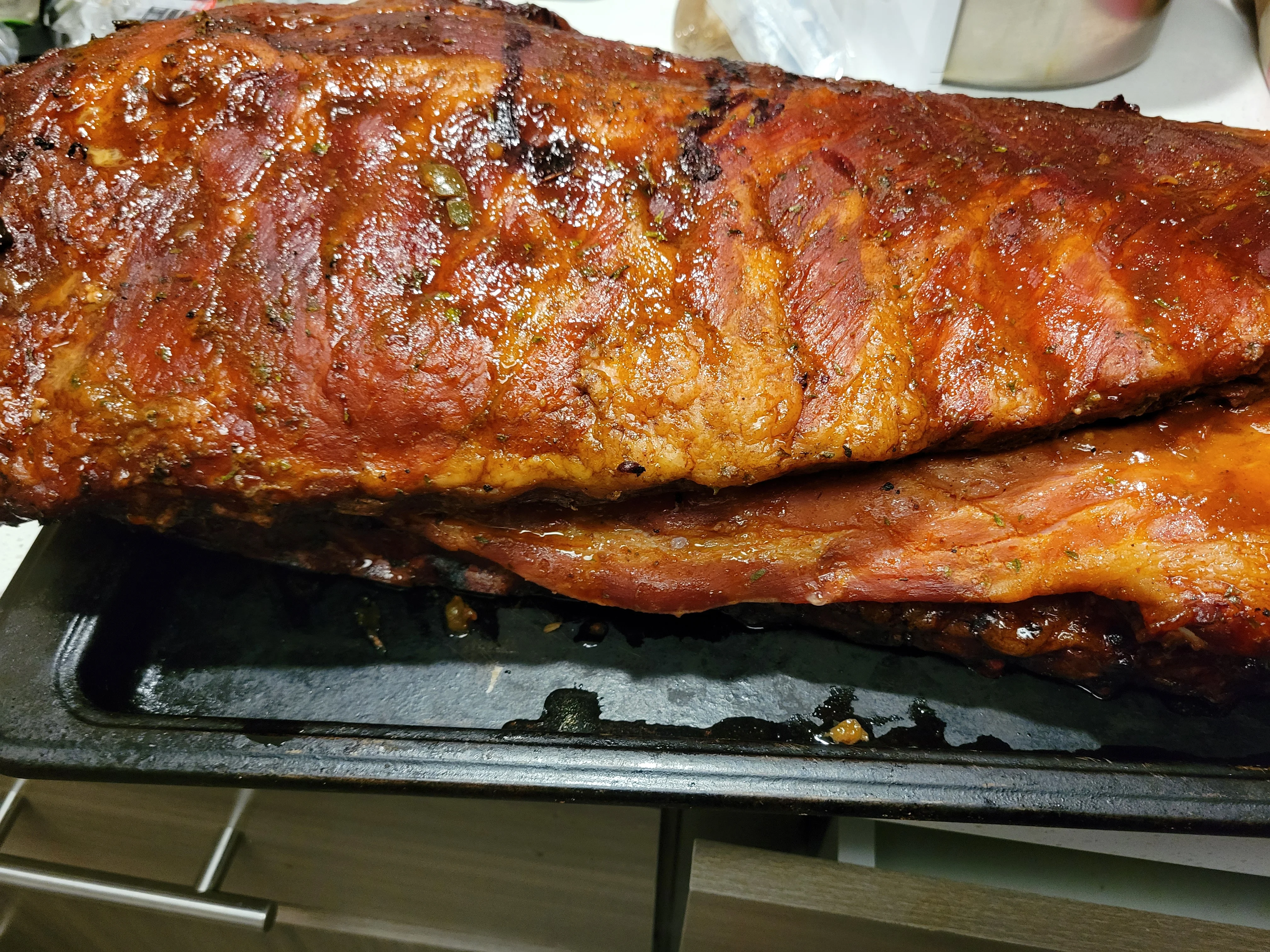 A large piece of glazed meat, likely ribs, resting in a dark tray. The meat has a caramelized coating and appears juicy.