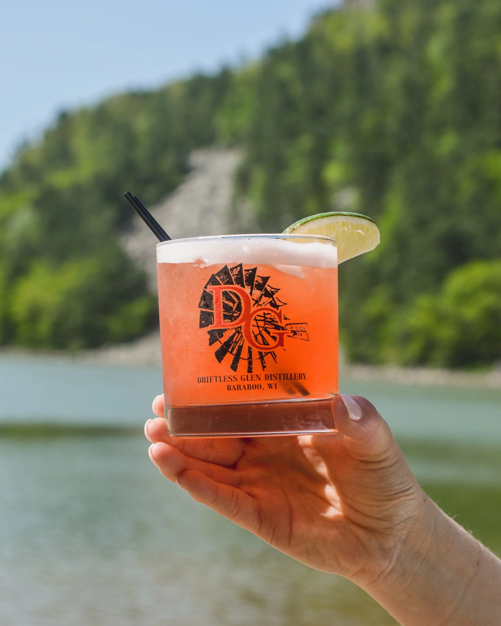 Beach Please cocktail in a pretty pink color being held up with water and sand and trees in the background 