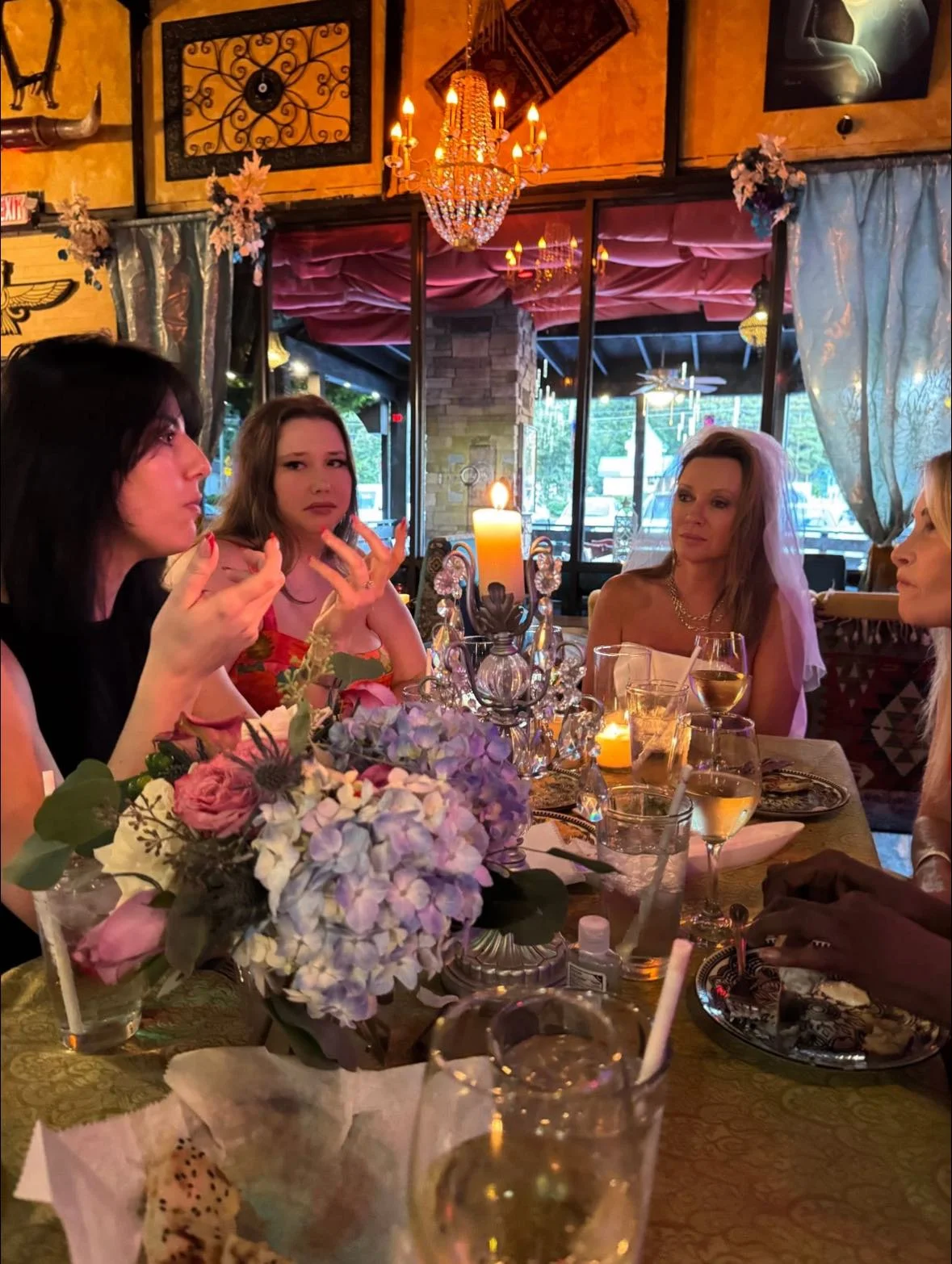 A dining scene with women engaged in conversation around a table adorned with flowers and candles, set against an ornate restaurant interior.