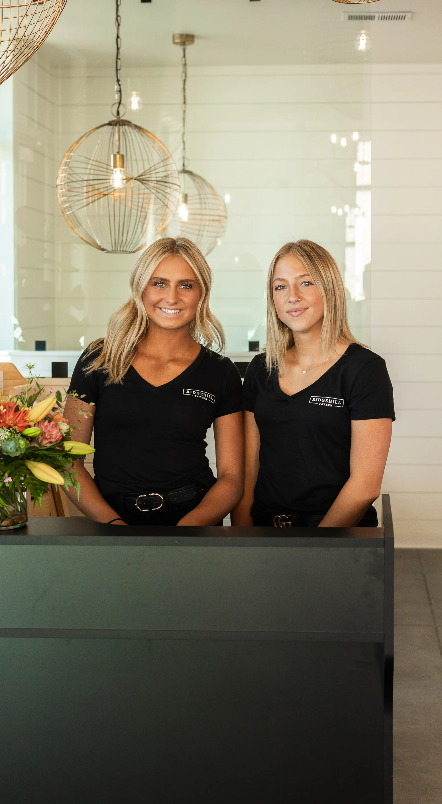 Two women at a restaurant reception desk with a floral arrangement.