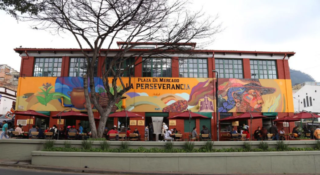 A vibrant market plaza with a large mural on the building's facade. Outdoor seating is visible, surrounded by trees.