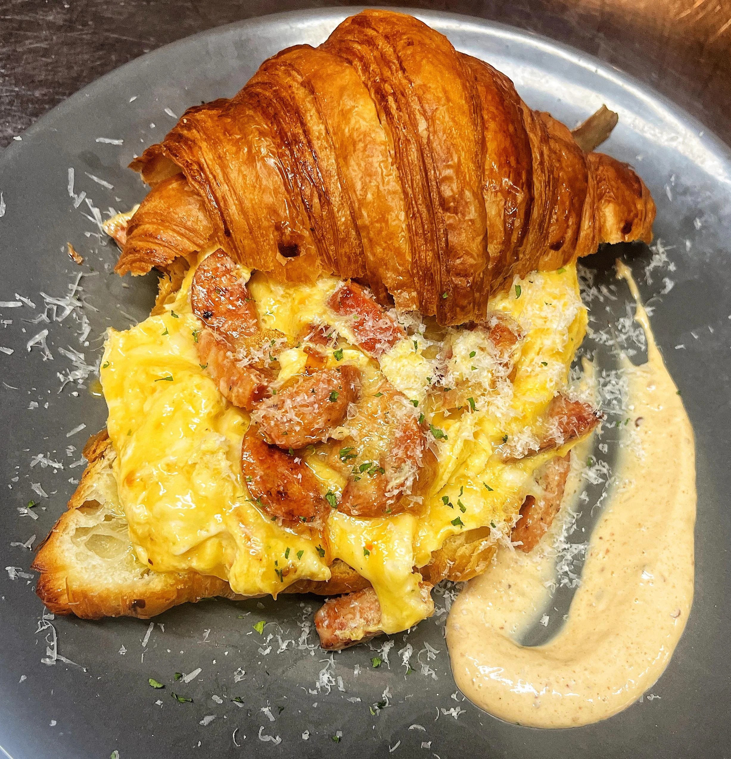 A croissant filled with scrambled eggs and shrimp, sprinkled with cheese, served with a creamy sauce on a dark plate.