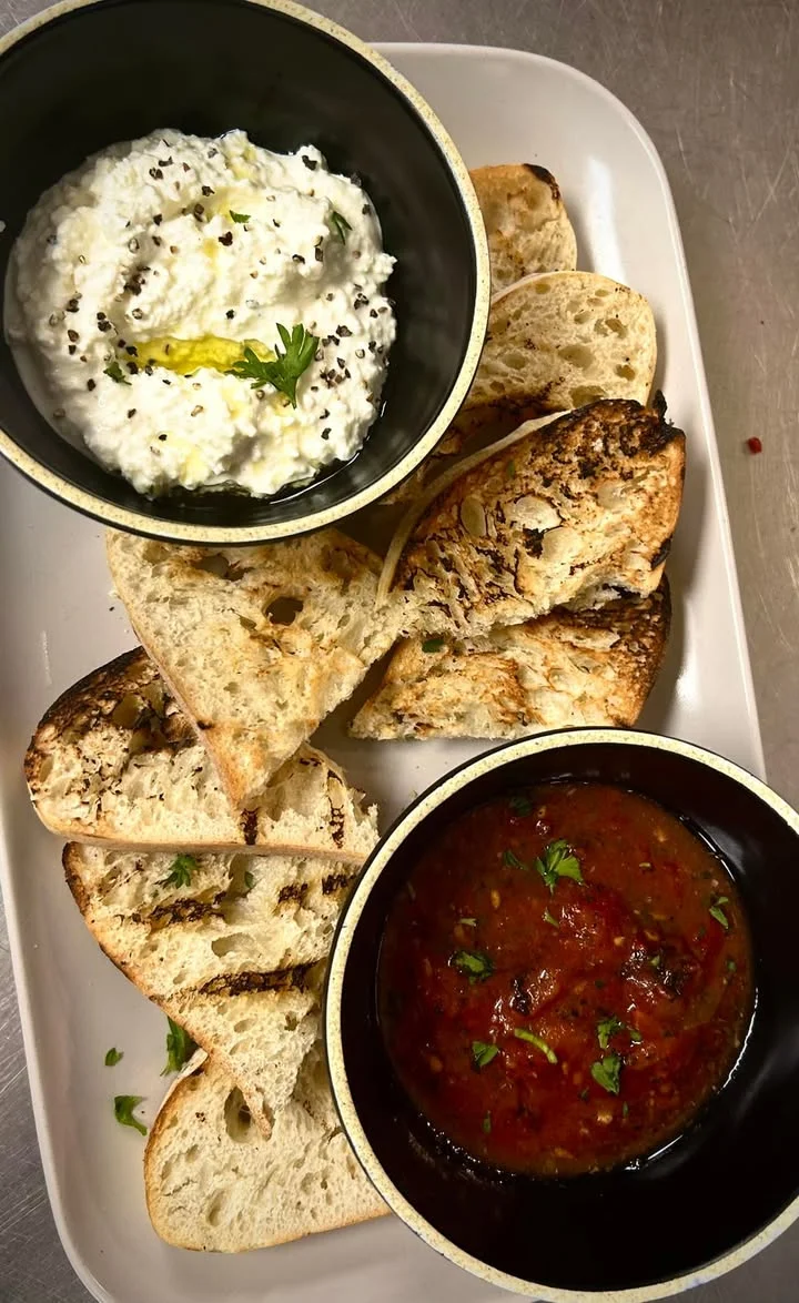 A plate featuring grilled bread accompanied by a bowl of creamy dip and a bowl of tomato-based sauce, garnished with herbs.