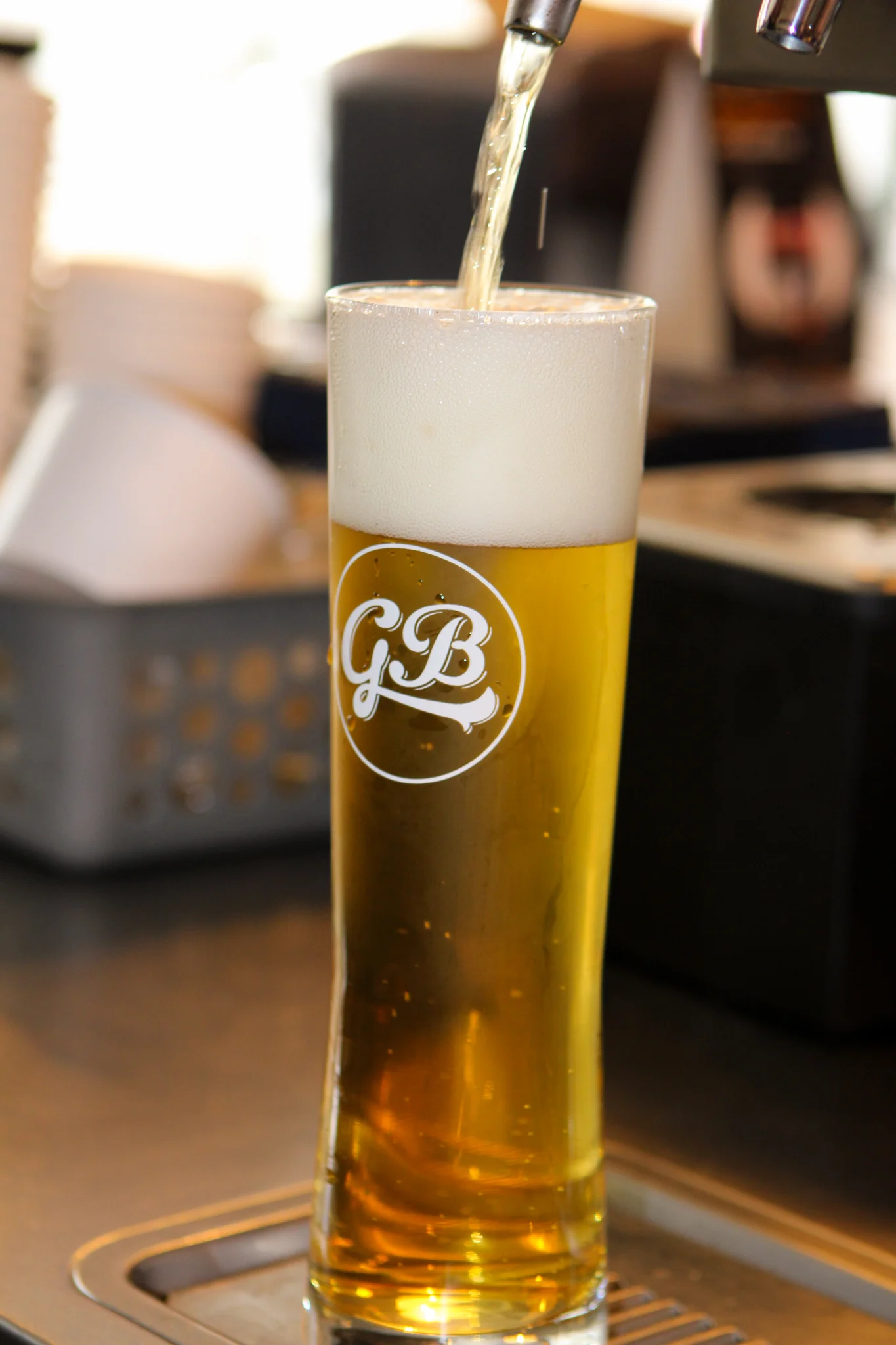 A glass beer mug being filled with golden beer and topped with foam. The glass features a logo, and the background includes blurred items.