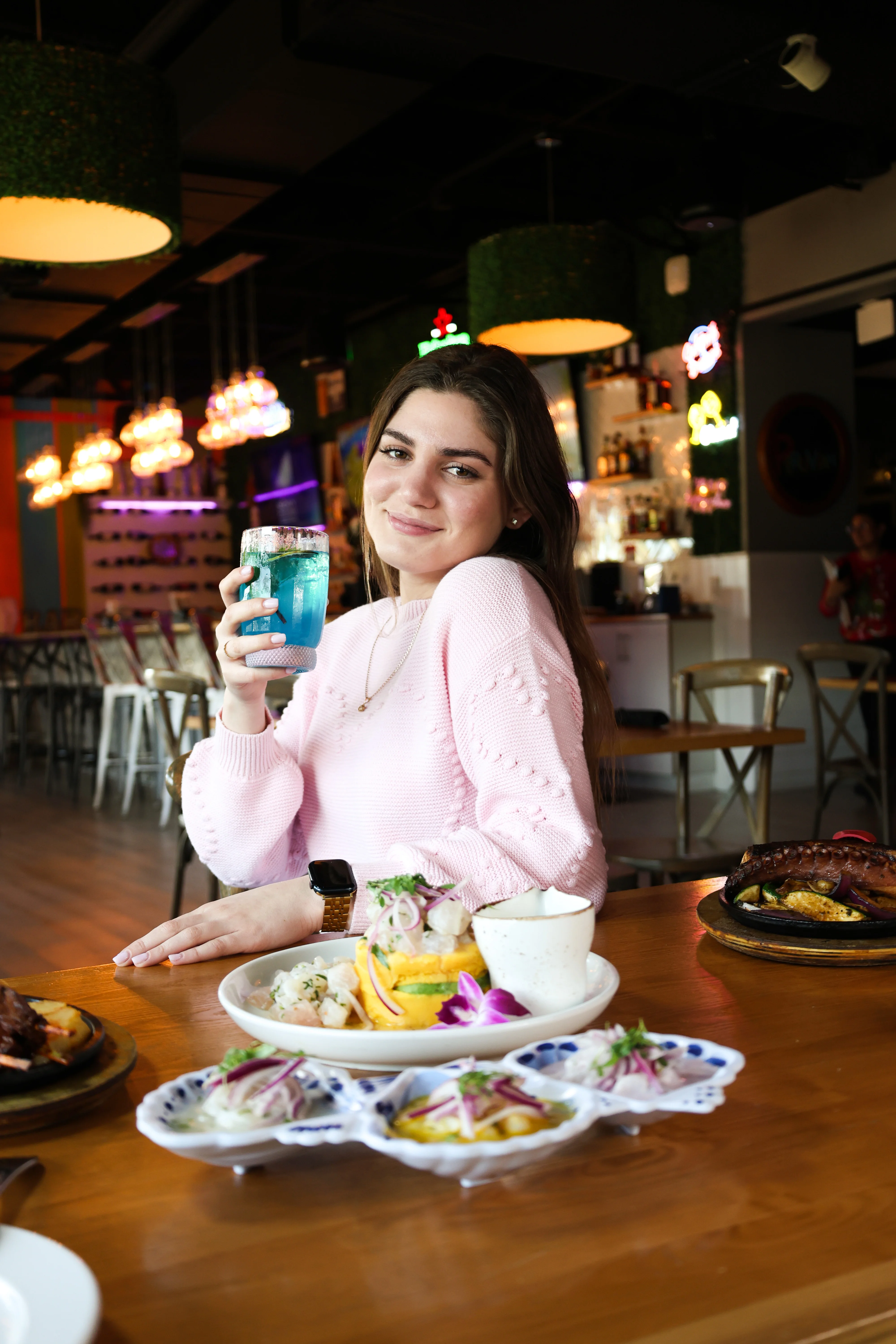 A young woman enjoying a colorful drink at a restaurant with delicious food.
