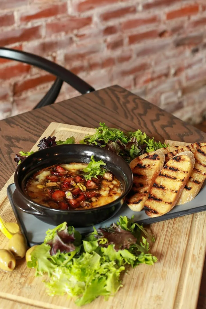 A bowl of melted cheese with toppings, served alongside grilled bread and fresh greens on a wooden cutting board.
