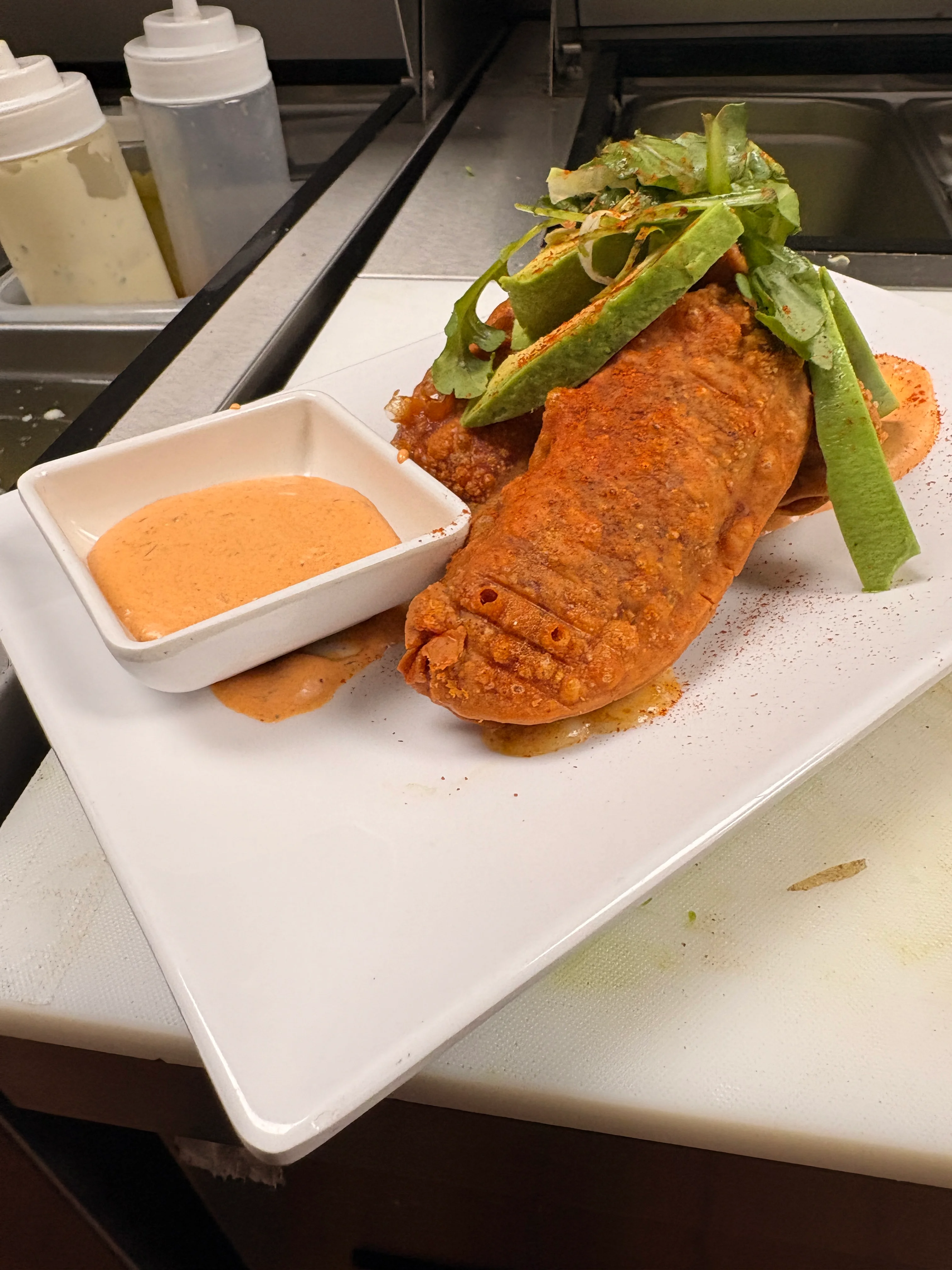 A plate featuring a crispy fried item topped with herbs and served with a side of dipping sauce. The dish is garnished with fresh greens and spices.