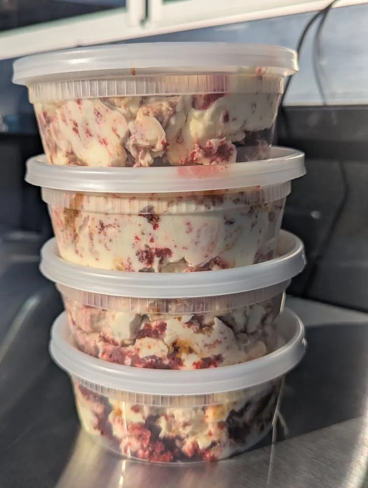 A takeout container filled with a variety of dishes including green vegetables, pasta, meat rolls, and a cornbread muffin.