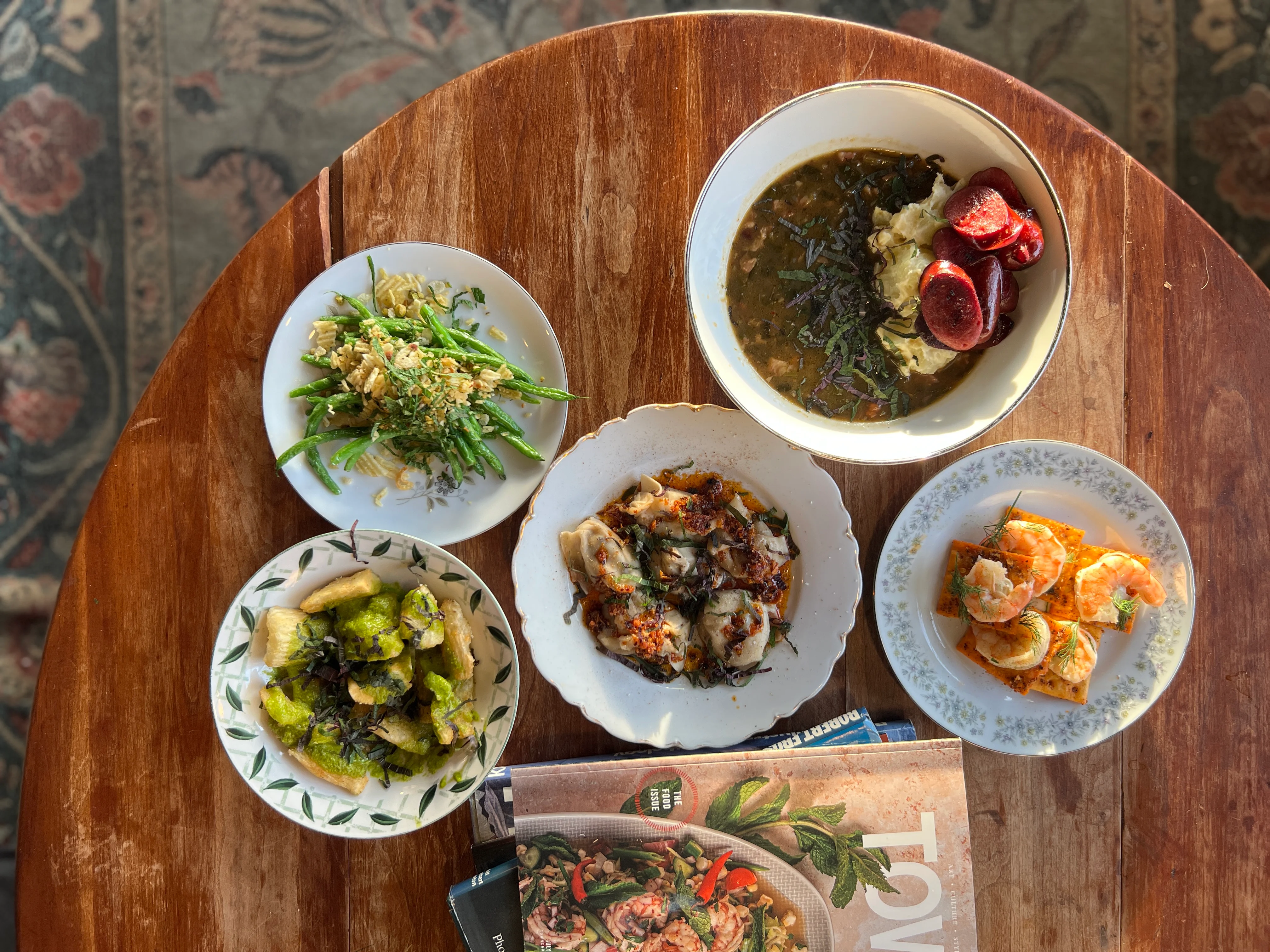 A variety of dishes arranged on a wooden table, including a bowl of soup, a plate of noodles with vegetables, and several small dishes with mixed ingredients.