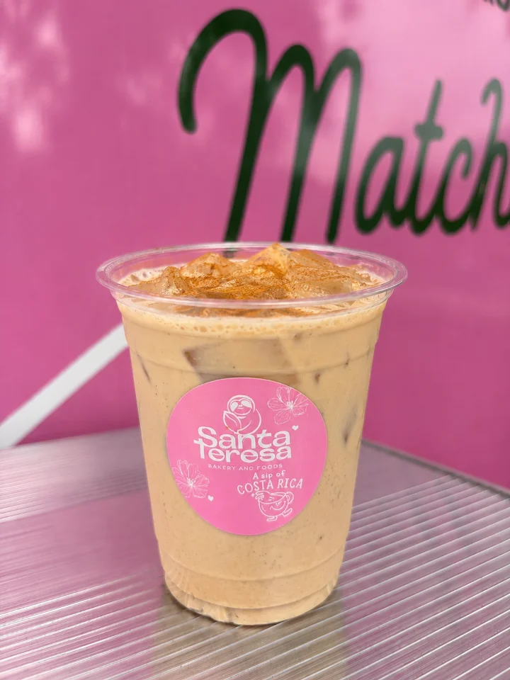 A clear cup filled with iced coffee topped with cinnamon, presented in front of a pink backdrop with coffee branding.