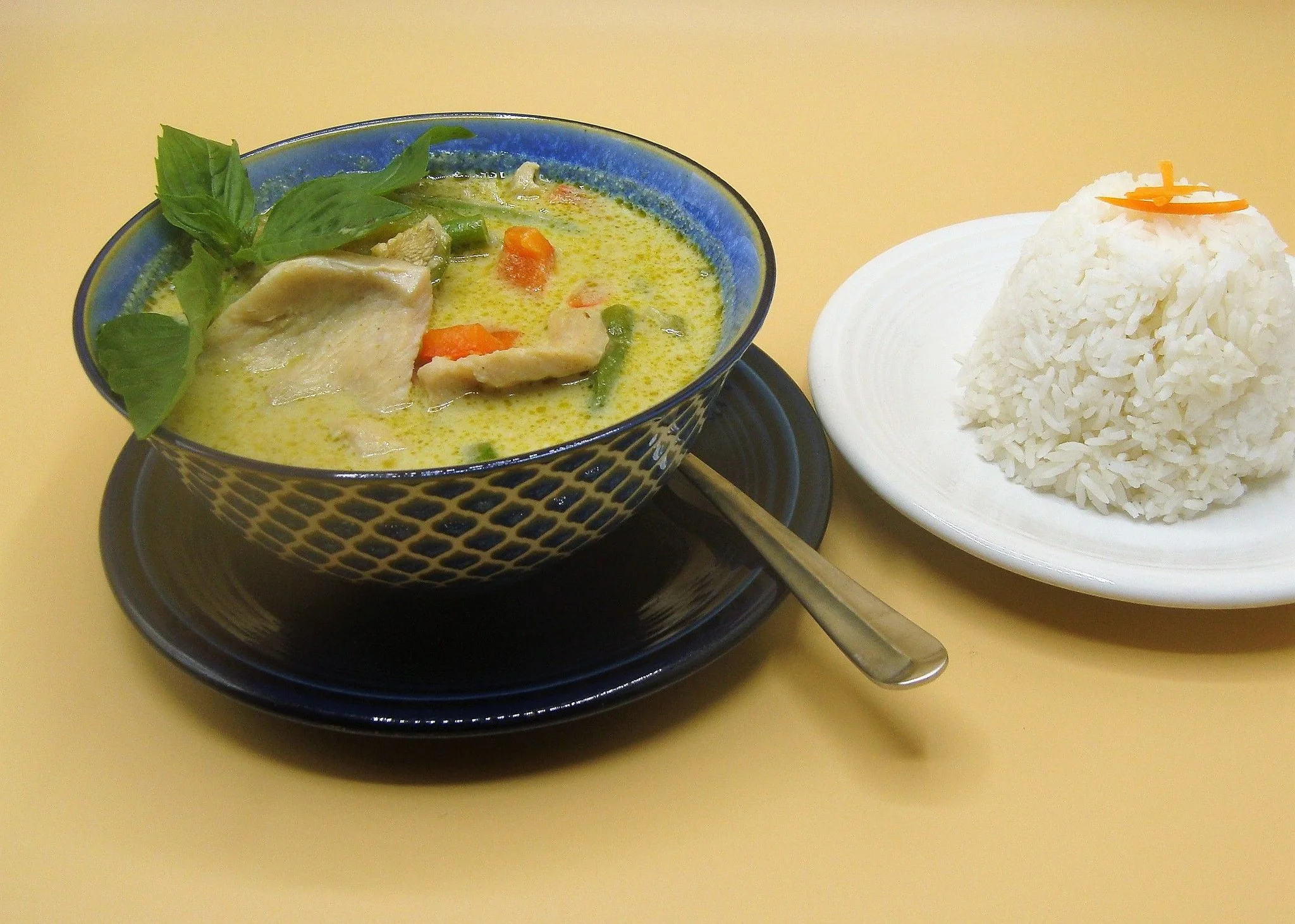 A bowl of creamy curry with assorted vegetables, served with a side of white rice and garnished with fresh herbs and lime wedges.