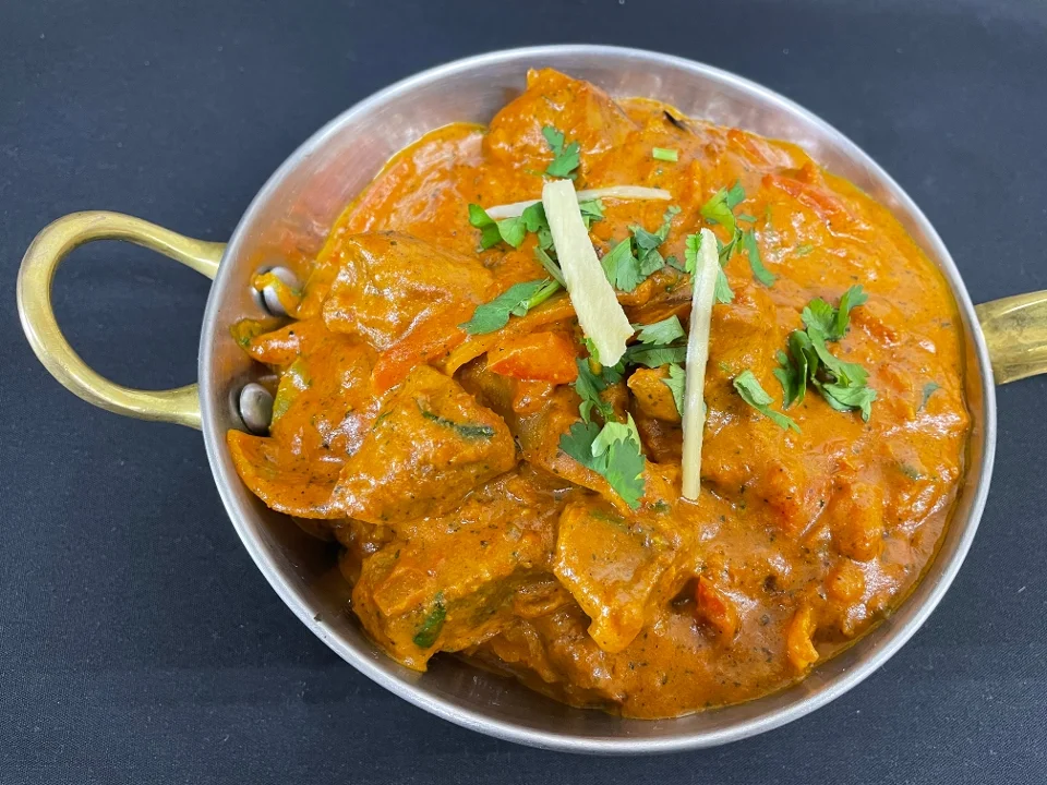 A metal dish filled with curry that includes chunks of meat and vegetables in a thick, orange gravy, topped with cilantro and ginger strips.