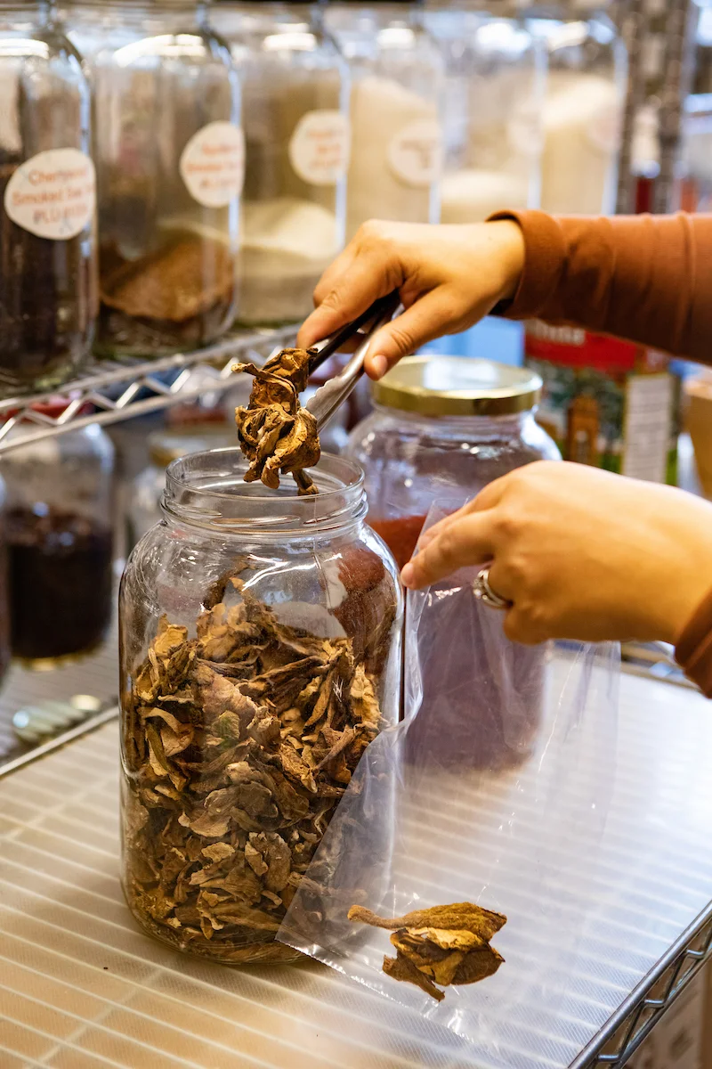 Person using tongs to select dried herbs from a glass jar