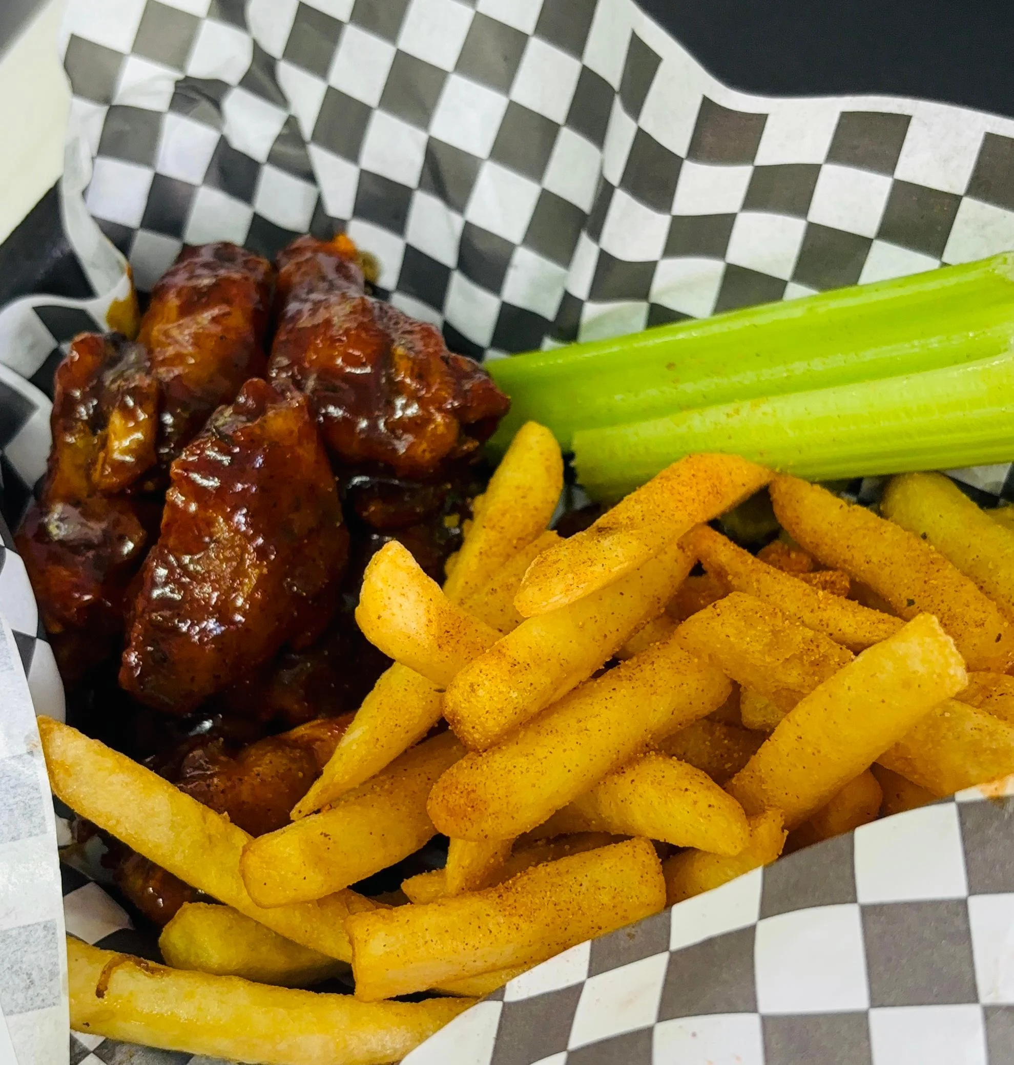 A serving of buffalo wings glazed with sauce, accompanied by crispy French fries and a stick of celery, all presented in a checkered paper-lined basket.