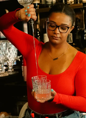 A bartender wearing glasses serves a colorful cocktail over ice. The background features a well-stocked bar with bottles.