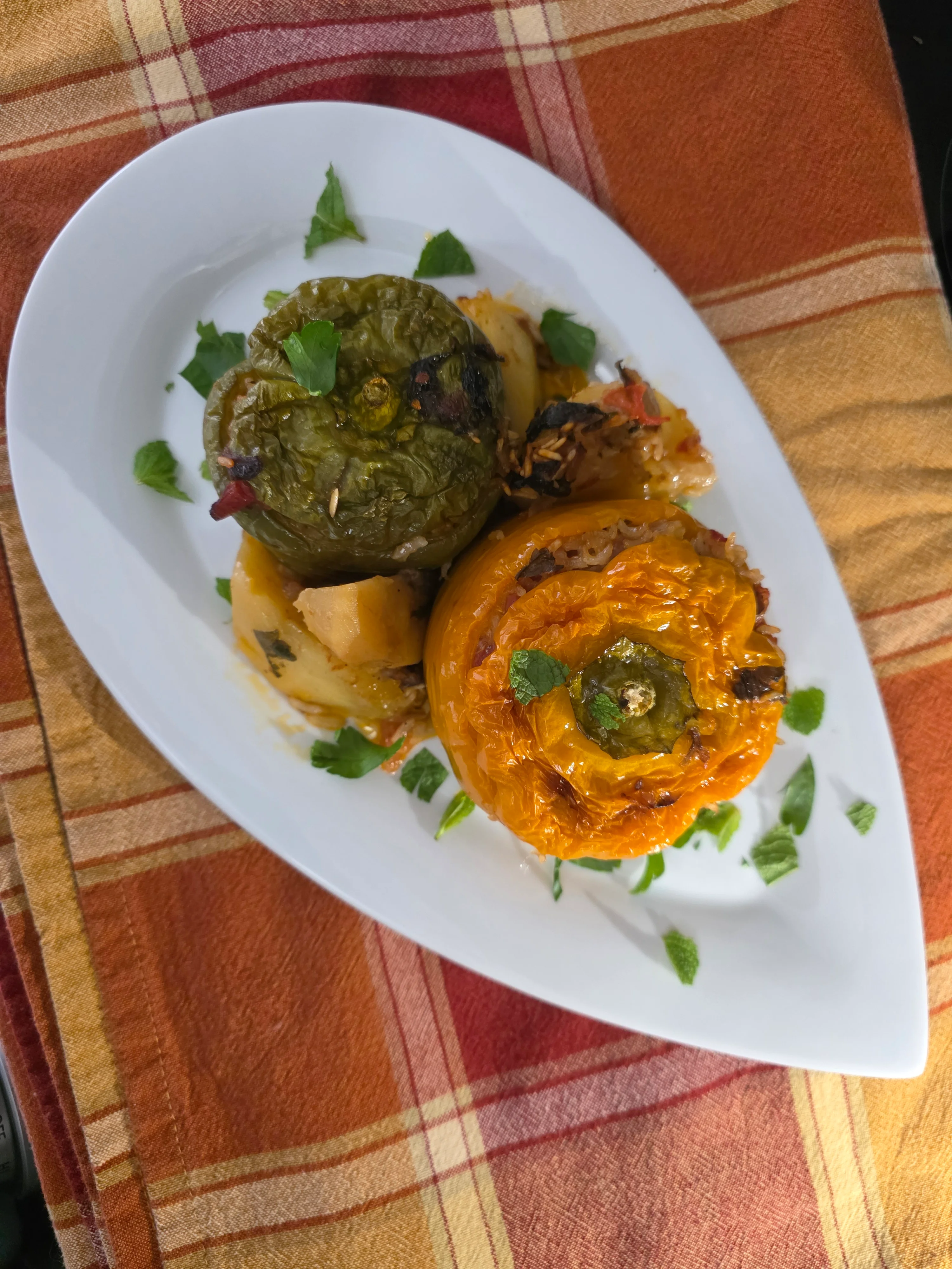 A serving of stuffed peppers, one green and one orange, placed on a white dish garnished with fresh herbs, surrounded by cooked vegetables.