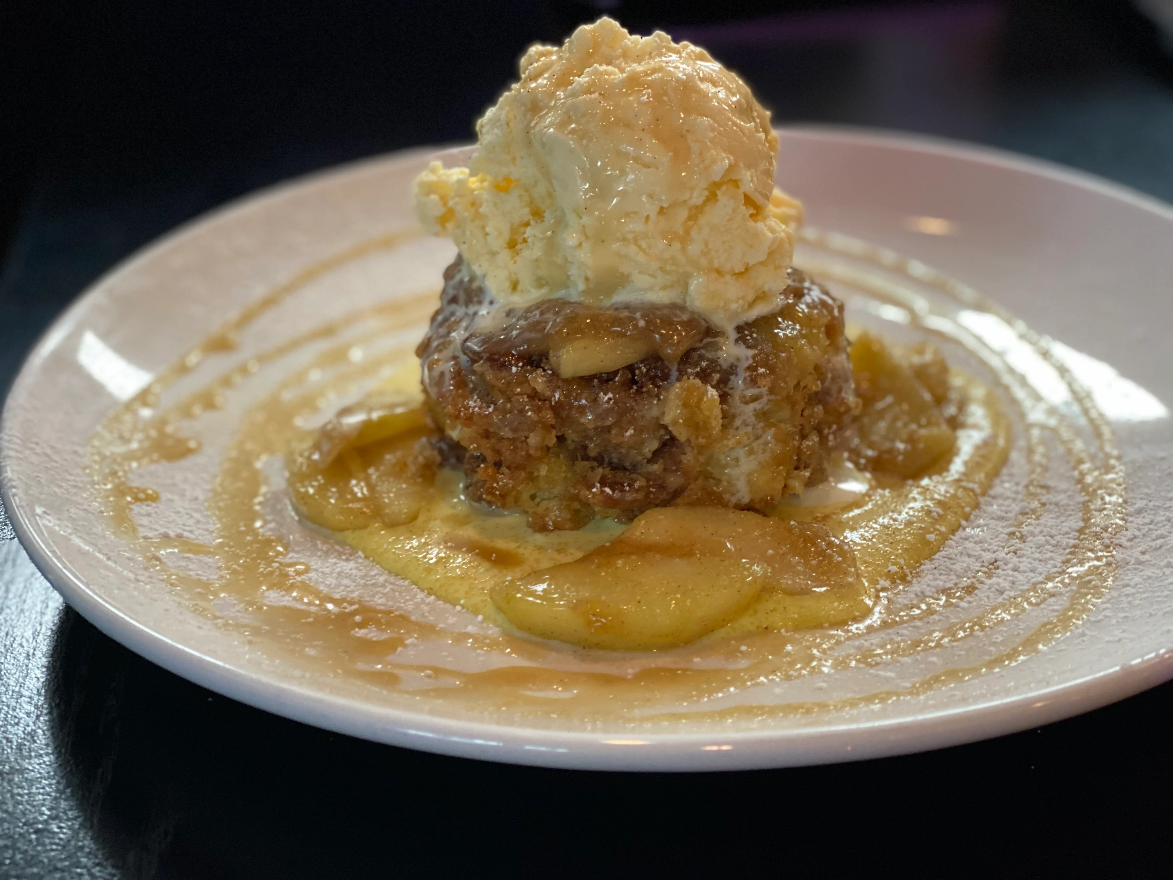 A warm dessert featuring a cake topped with melted apples and a scoop of vanilla ice cream, drizzled with a sweet sauce.