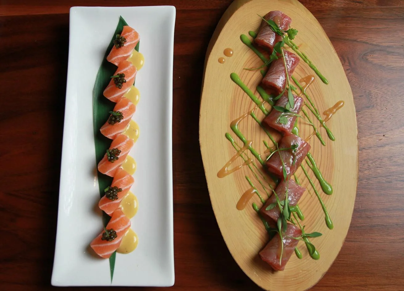 An overhead shot of two elegant appetizer platters on a dark wooden table: a long white rectangular plate on the left with a zigzag arrangement of fresh salmon rolls topped with caviar and microgreens over a yellow sauce, and an oval wooden board on the right displaying raw tuna carpaccio rolls garnished with microgreens and drizzled with green and orange sauces.